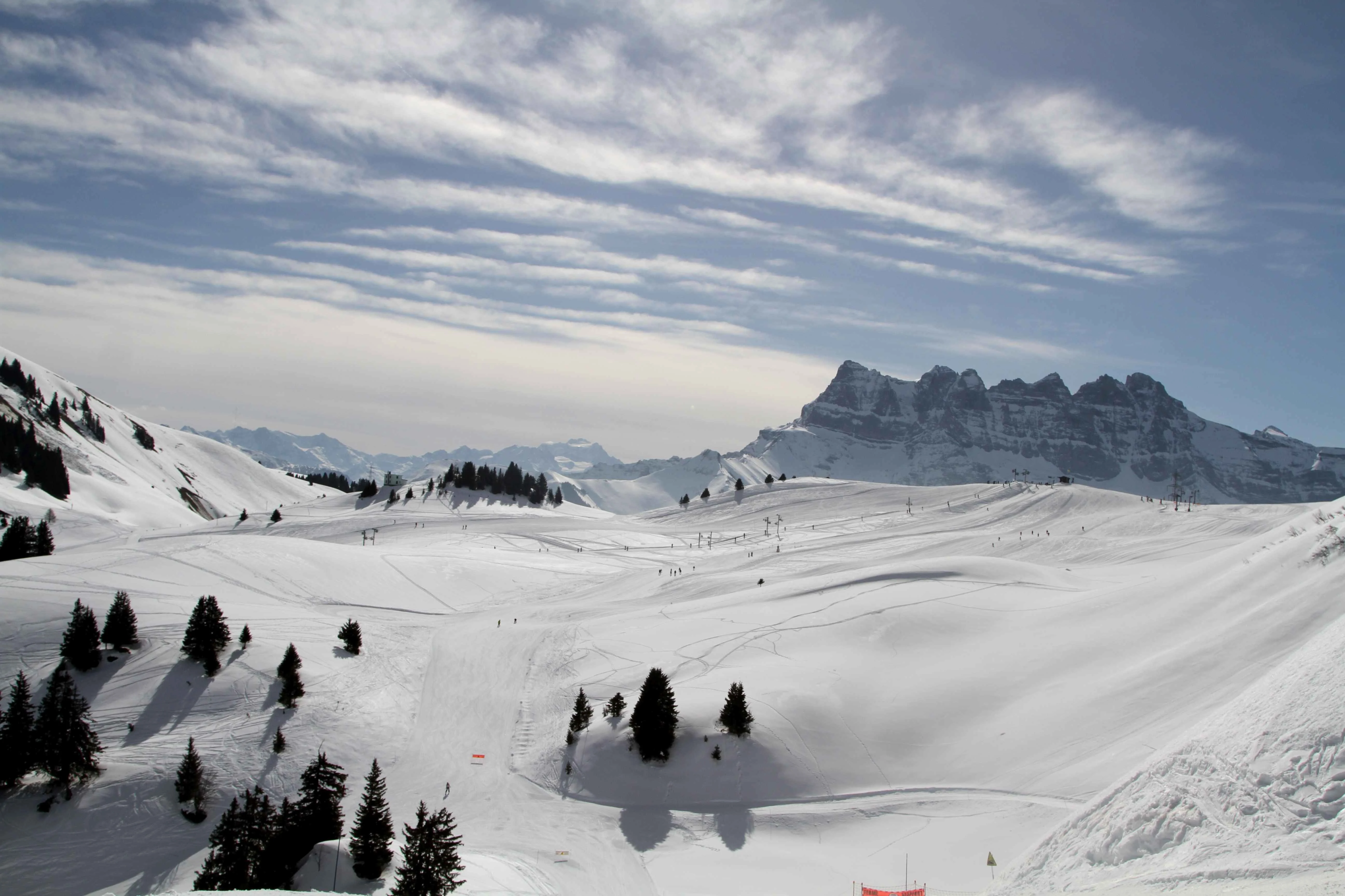 Snow-covered slopes and scattered evergreens in Châtel with the Dents du Midi mountains in the background under a blue, partly cloudy sky.
