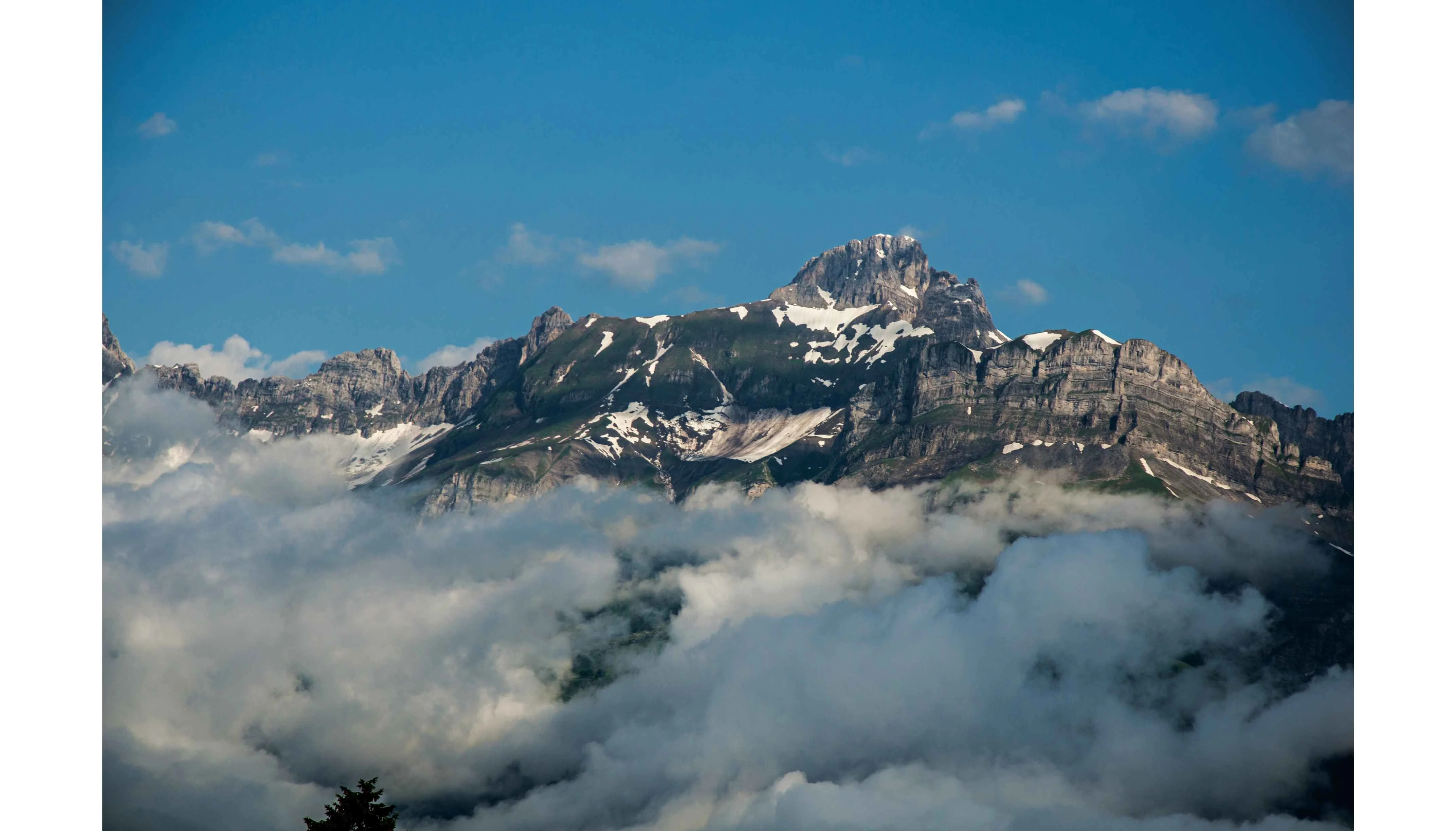 Dramatic snow dusted mountain peak emerging above clouds under a blue sky near Saint-Gervais-les-Bains, French Alps.