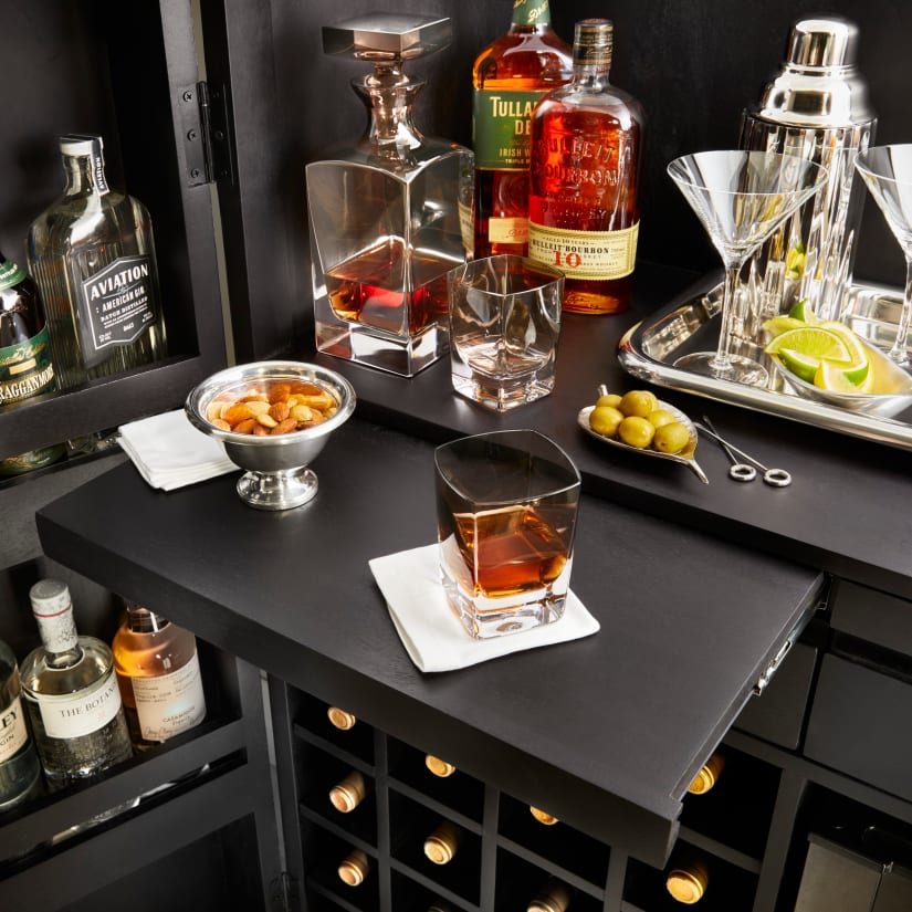 Bar counter setup with cocktail shaker, glassware, limes, and a big square ice decanter on a black surface.