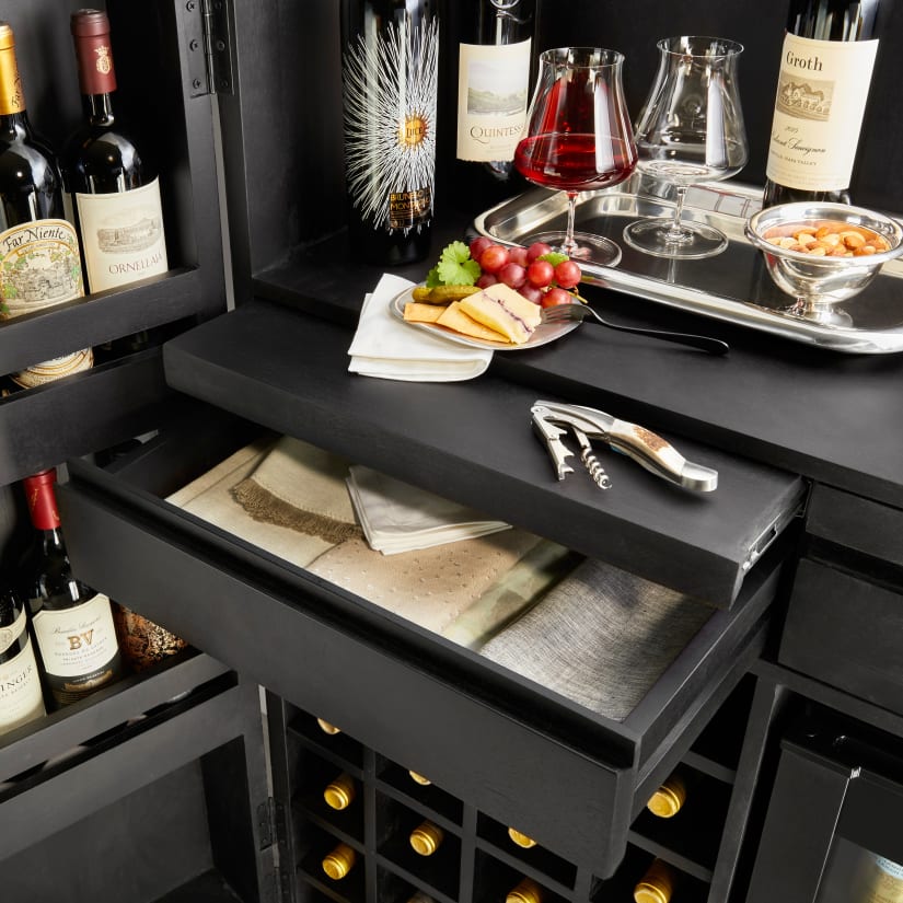 Cluttered home bar interior with wine bottles, a drawer open showing a towel and napkin, and a drawer pull.