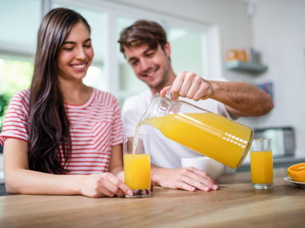 Bir çiftin önünde portakallar ve portakal suyu dolu bir sürahi var, erkek olan sürahiden bardağı c vitamini kaynağı olan portakal suyu koyuyor