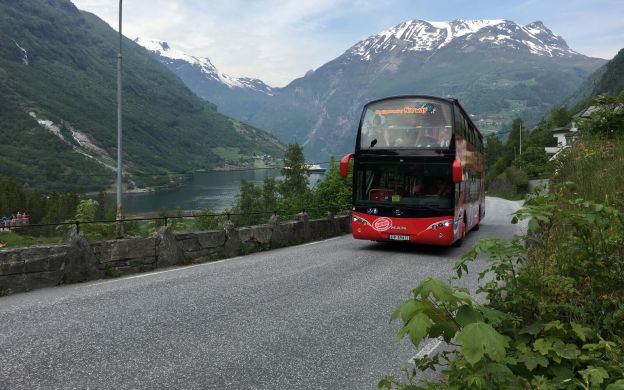 A striking red hop-on hop-off bus cruising through the streets of Geiranger, framed by majestic mountains, creating a stunning scenic experience.