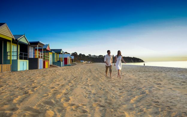 A couple relishing their time at a Melbourne beach, with colorful hut-shaped houses dotting the shoreline in the background.
