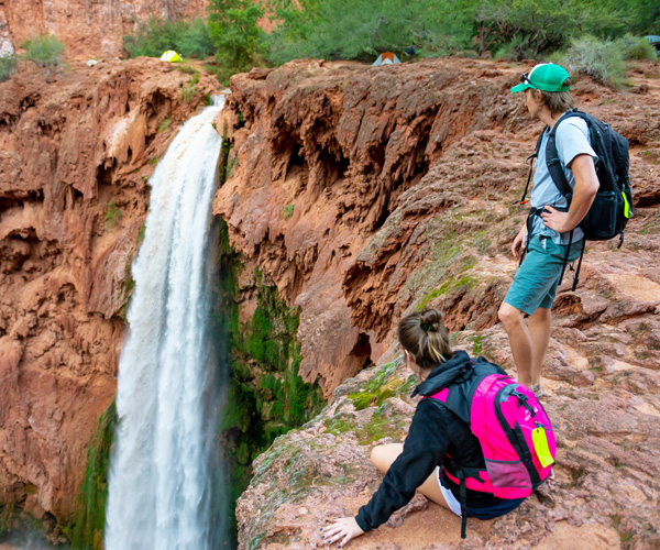 A man and woman stand at the edge of a cliff overlooking a waterfall. Tents and trees can be seen in the background