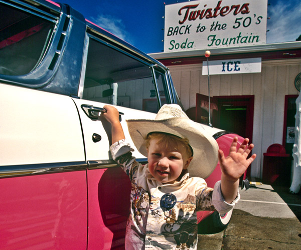 A little boy in a cowboy costume stands next to a pink and white vintage car in front of Twisters restaurant