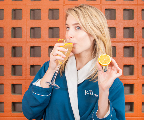 A blonde girl in a blue bathrobe drinks an orange cocktail