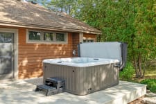 A hot tub on a wooden deck beside a house, surrounded by trees.
