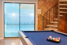 Interior view of a game room with a pool table, colorful billiard balls, and an indoor swimming pool visible through sliding glass doors.