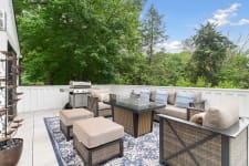 A contemporary patio setup with a grill and comfortable seating, framed by trees.