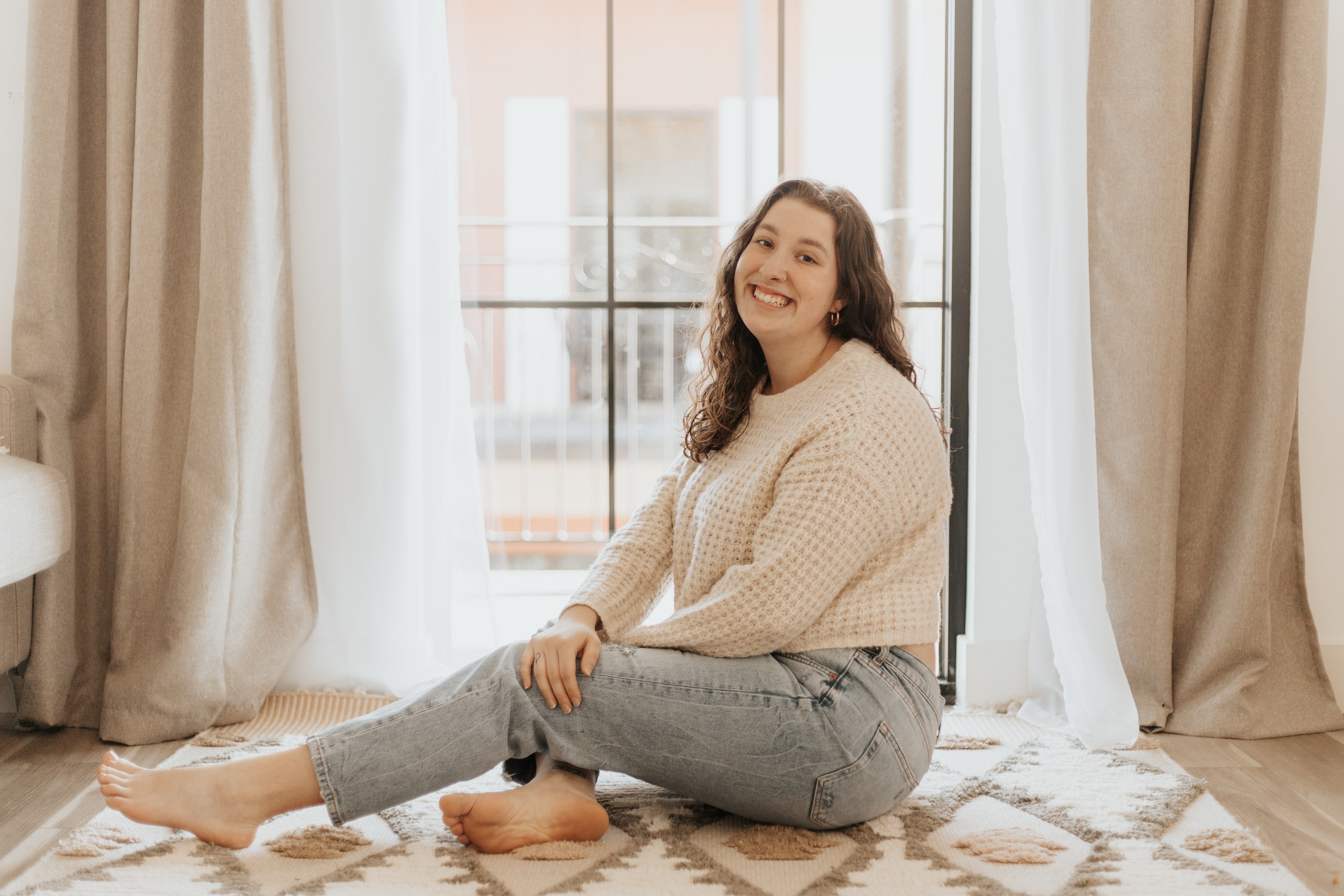 A young woman sitting on a rug in a sunlit room, smiling