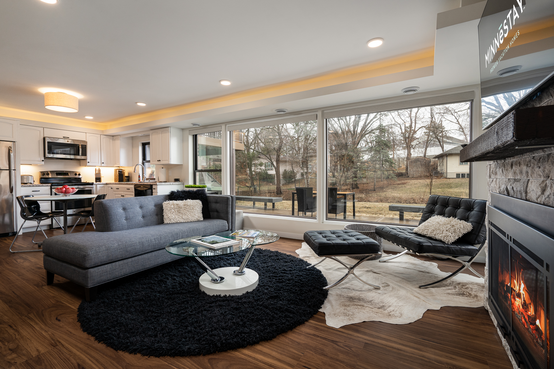 A modern living area with kitchen, featuring big windows and cozy decor.