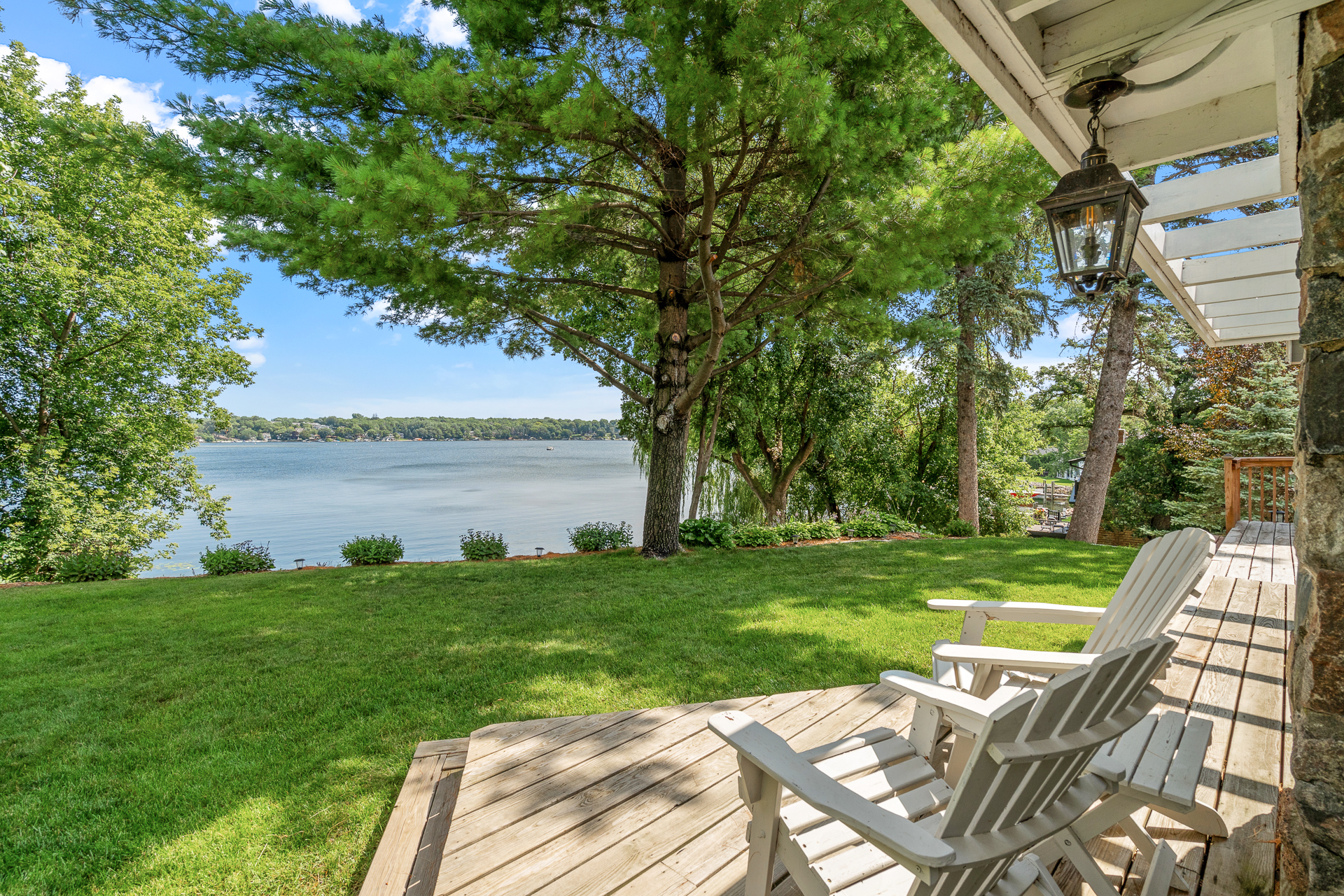 Lakeside view from a wooden deck with white adirondack chairs and green lawn.