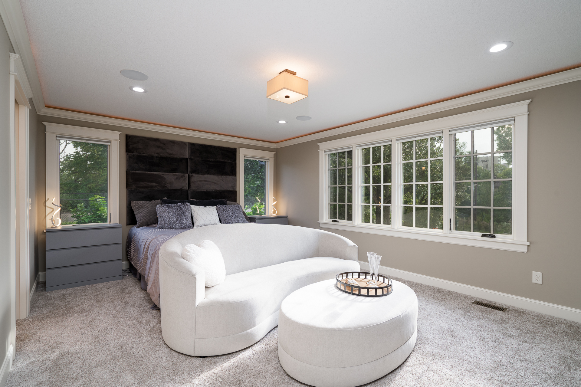 A modern and serene bedroom featuring a stylish bed, loveseat, and large windows.