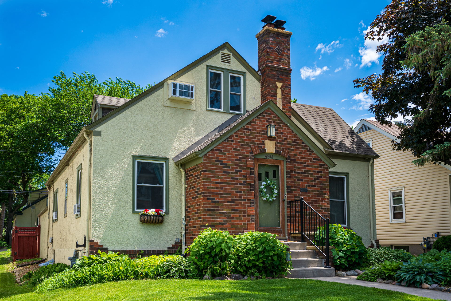 A charming residence with a mix of yellow stucco and red brick, featuring lush greenery and decorative accents.