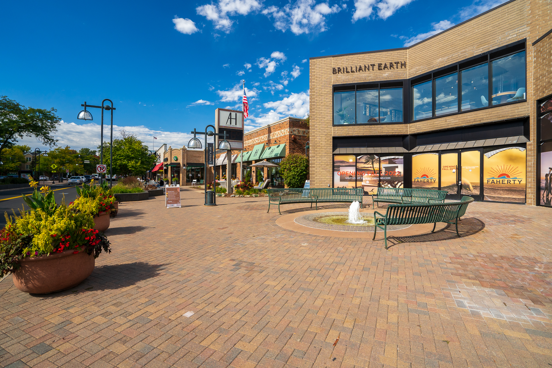 A sunny outdoor shopping plaza with a decorative fountain and vibrant storefronts.