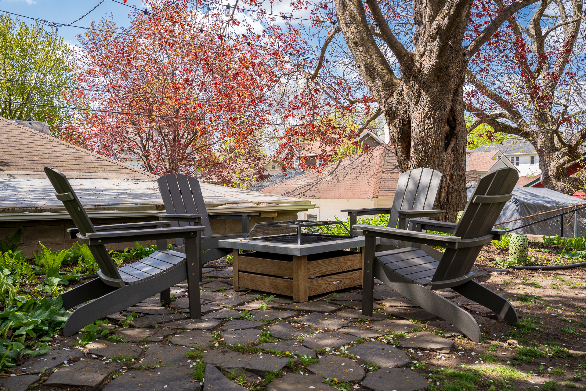 A quaint outdoor seating area with Adirondack chairs and a fire pit surrounded by greenery and spring foliage.