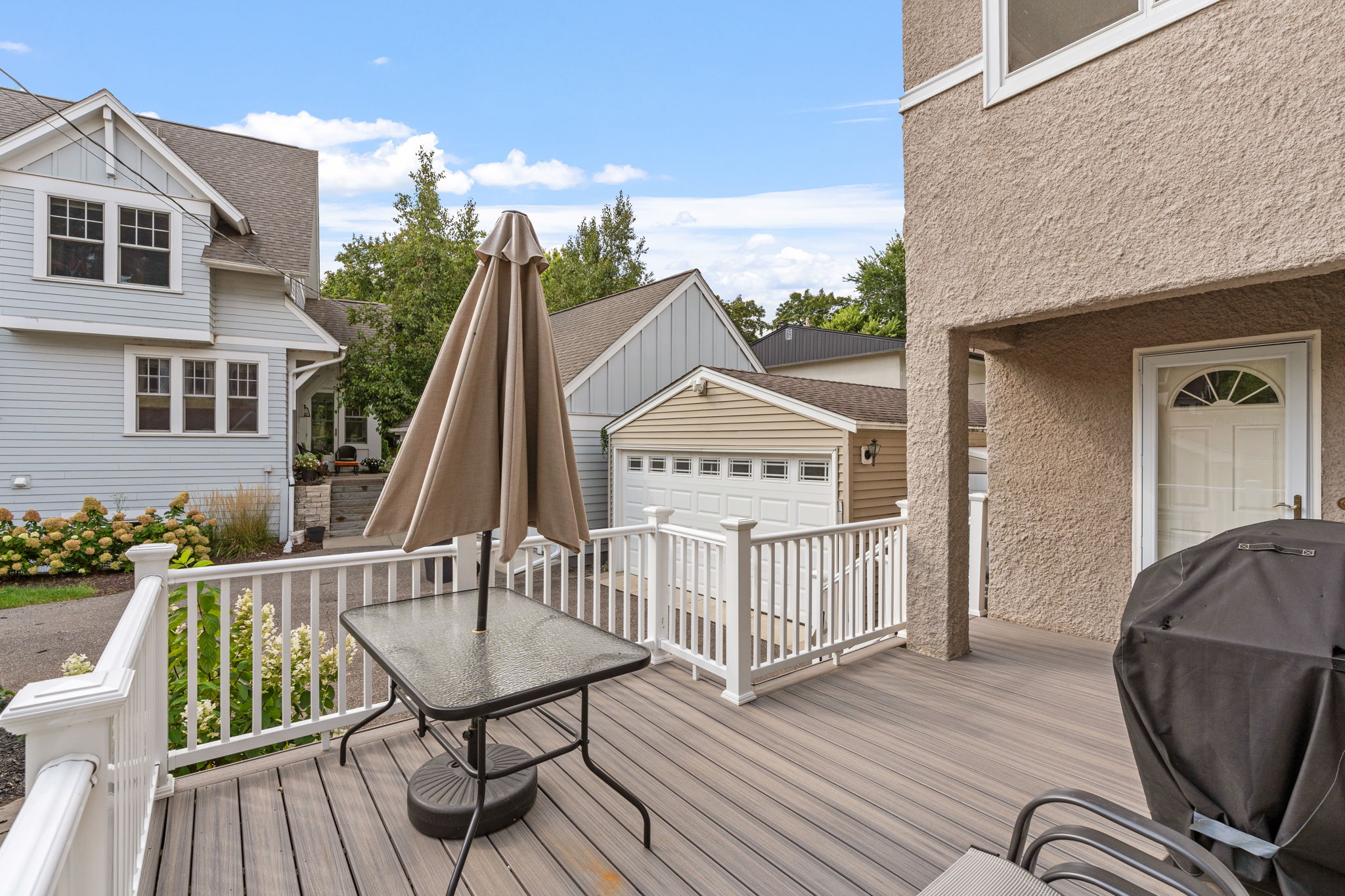 A peaceful outdoor deck with a glass table and umbrella, surrounded by charming homes and green scenery.