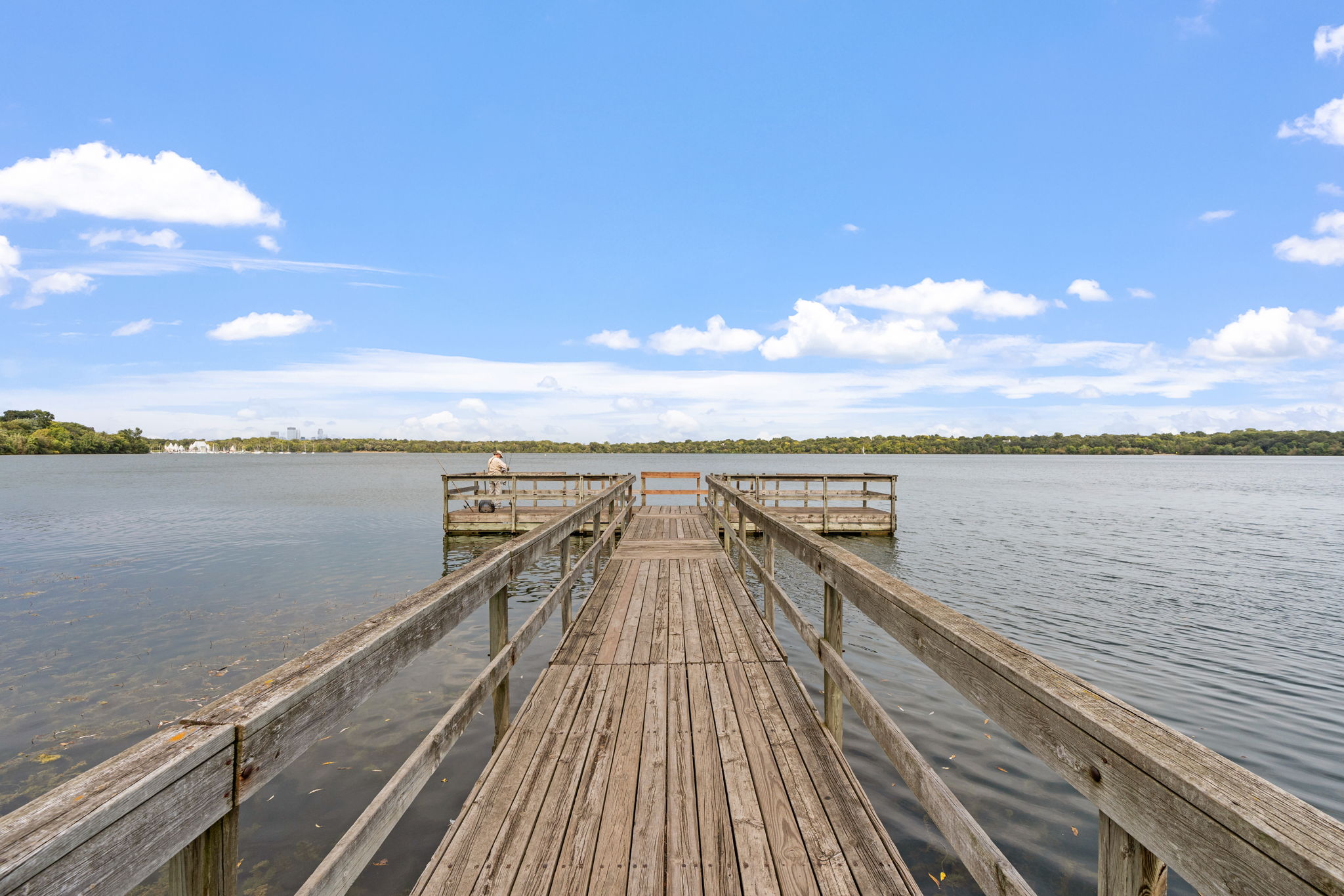 A tranquil wooden pier stretching into a serene lake under a blue sky with fluffy clouds.