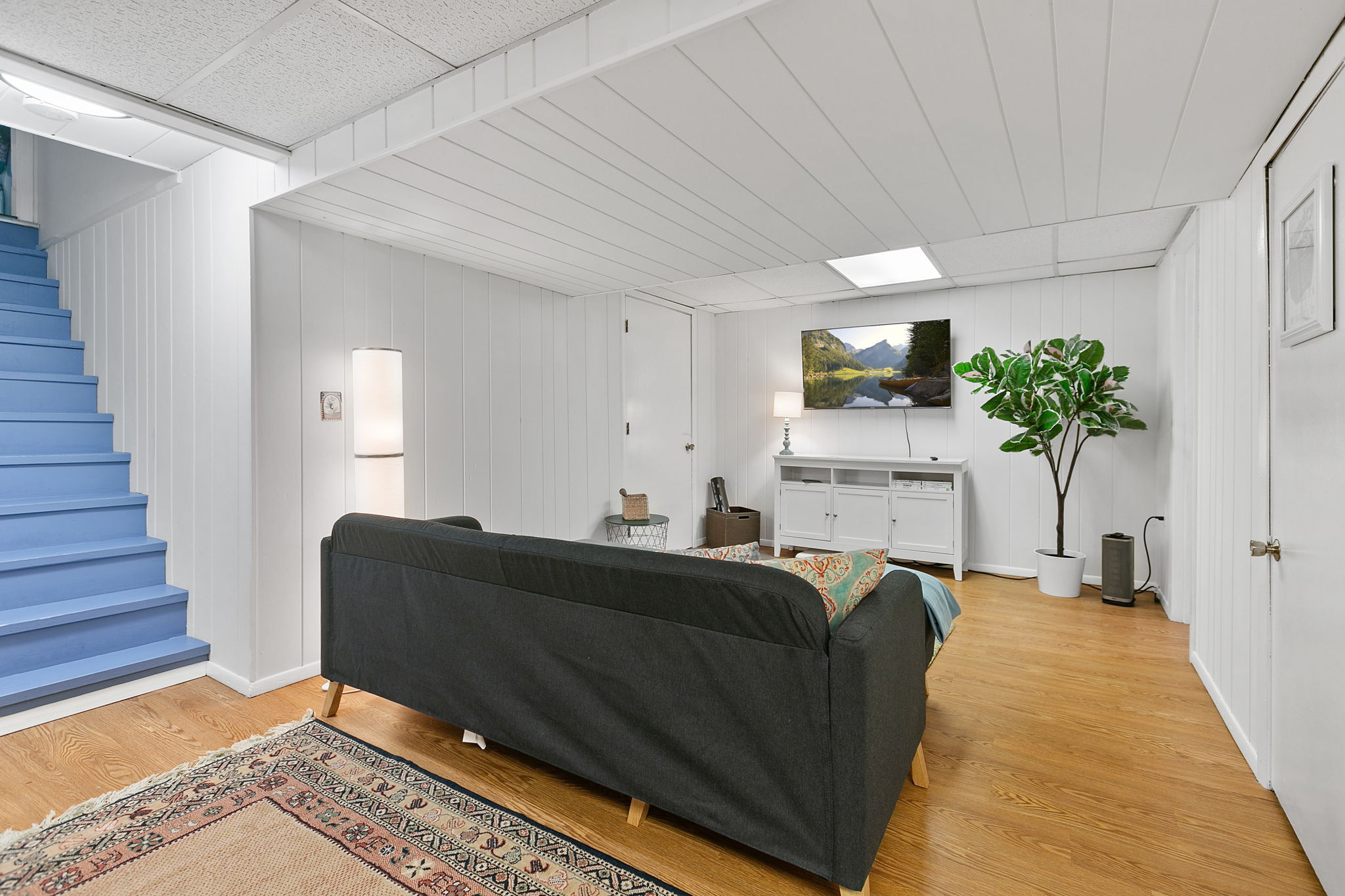 A cozy modern basement living area with a dark sofa, wall-mounted TV, and vibrant blue stairs.
