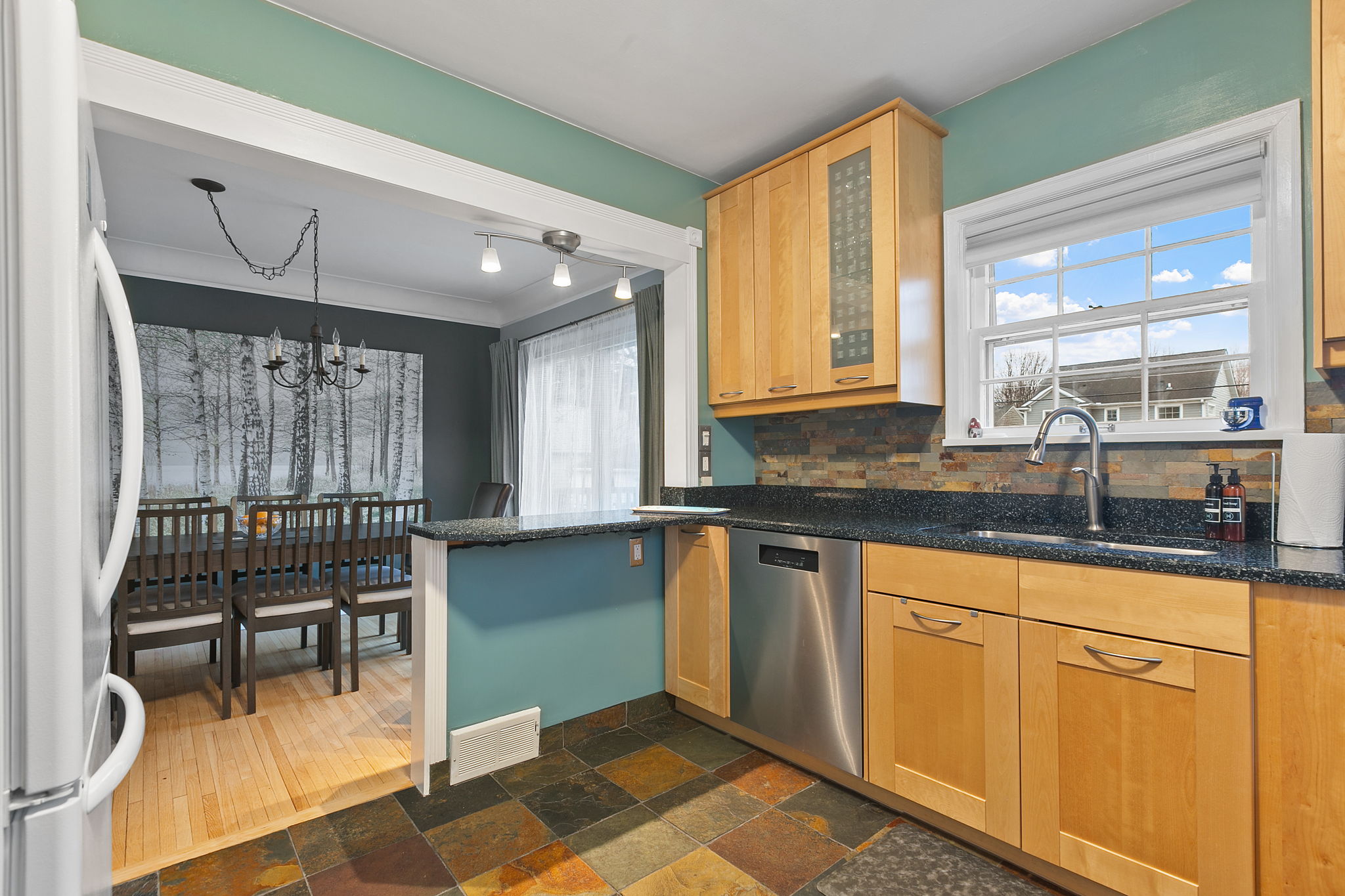 A modern kitchen with light wood cabinets and granite countertops, blending into a dining area with a forest painting.