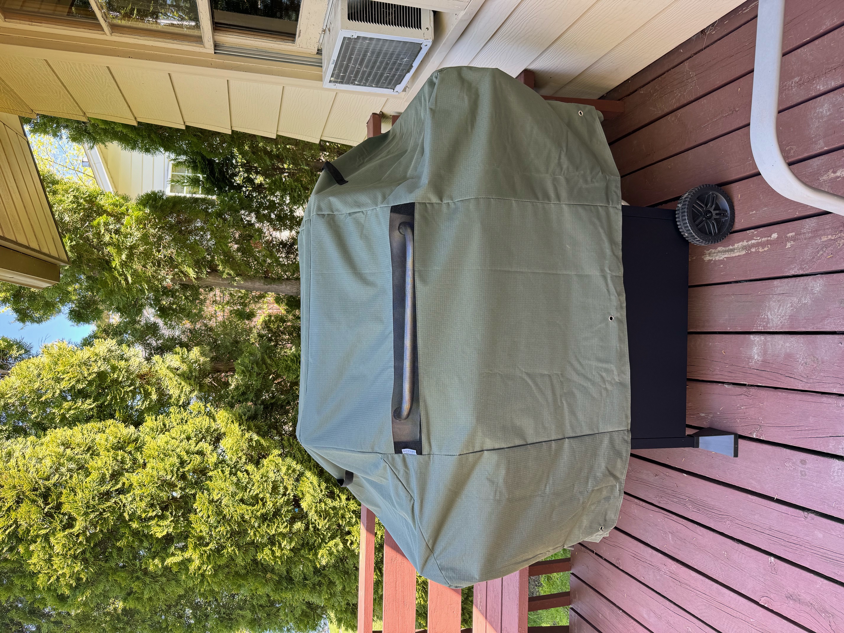 A covered grill on a sunny wooden deck surrounded by greenery.