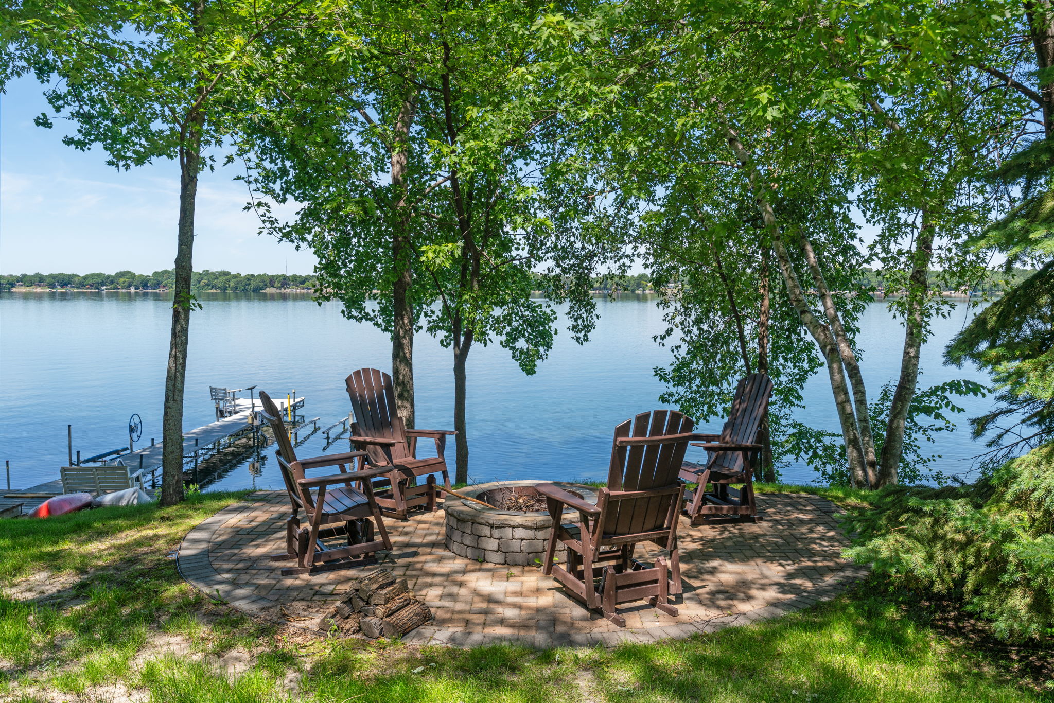 A peaceful lakeside fire pit area surrounded by Adirondack chairs under leafy trees.