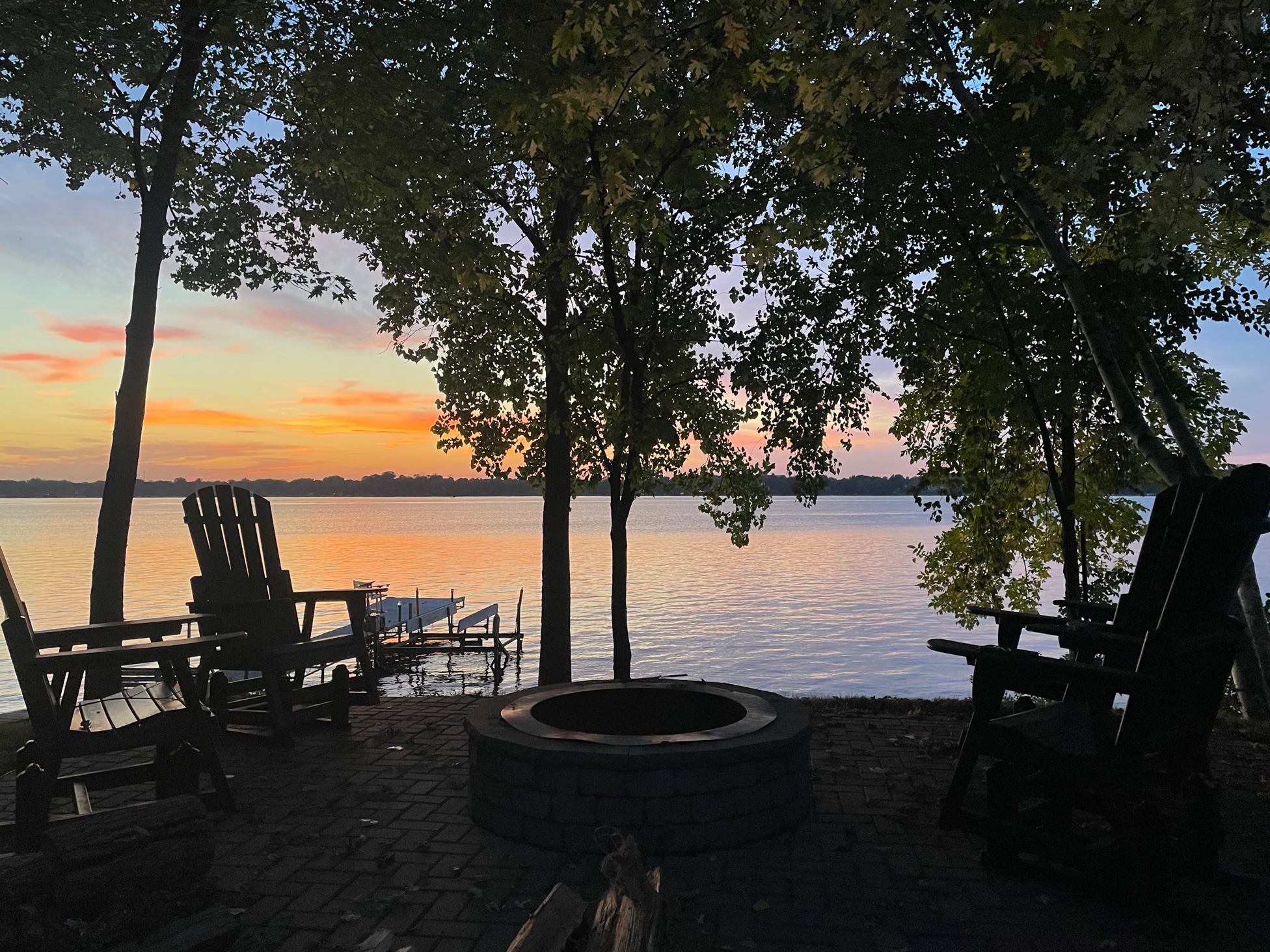 A peaceful lakeside scene at sunset featuring a fire pit and wooden chairs.