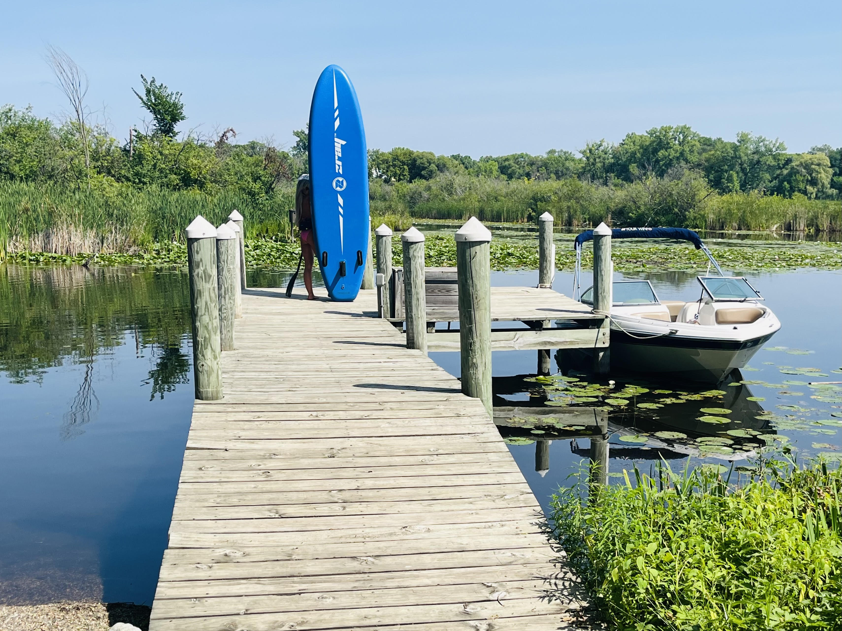 A person carries a blue paddleboard along a wooden dock by a calm lake.
