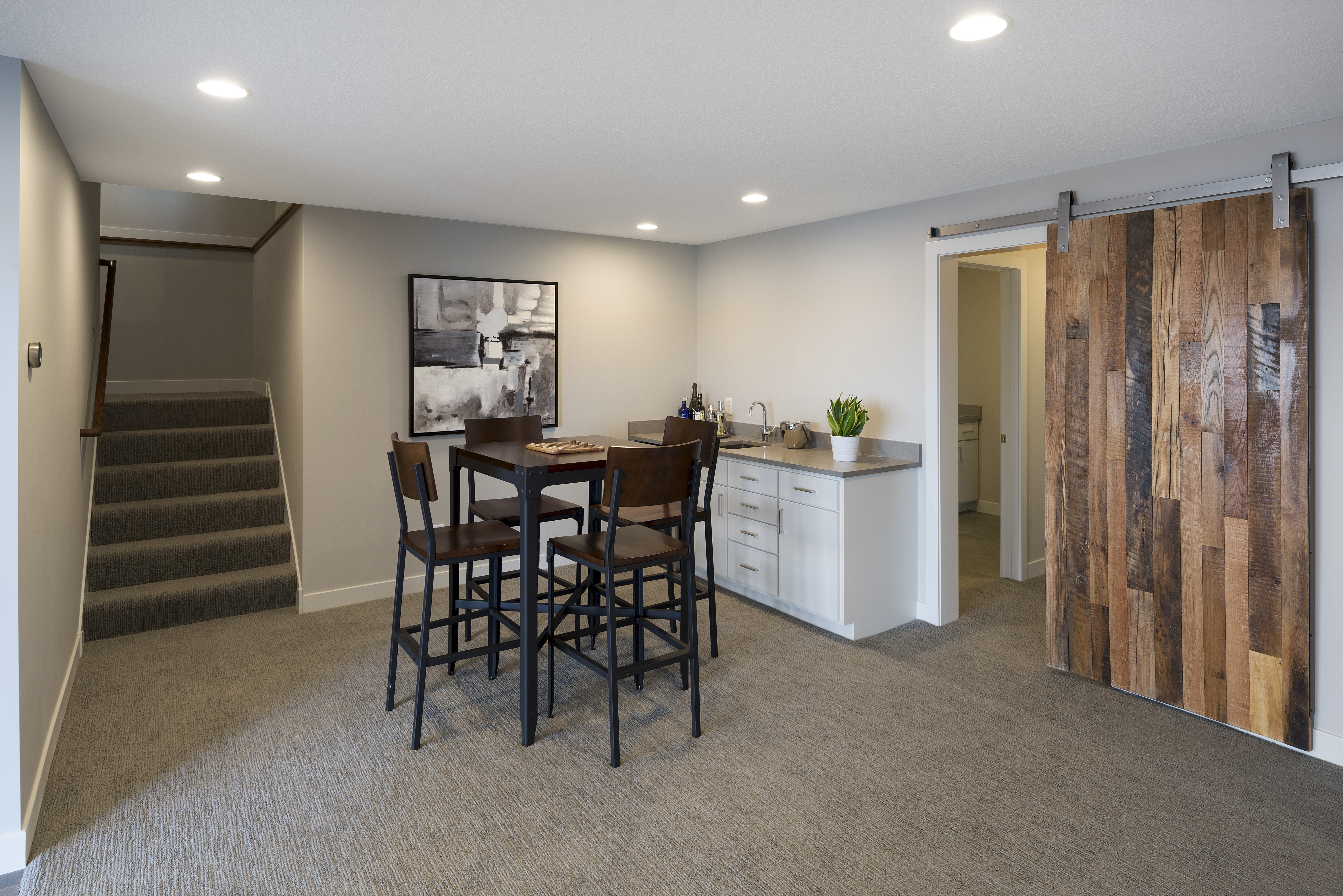 A modern basement living area with a dining set, kitchen space, and a sliding barn door.