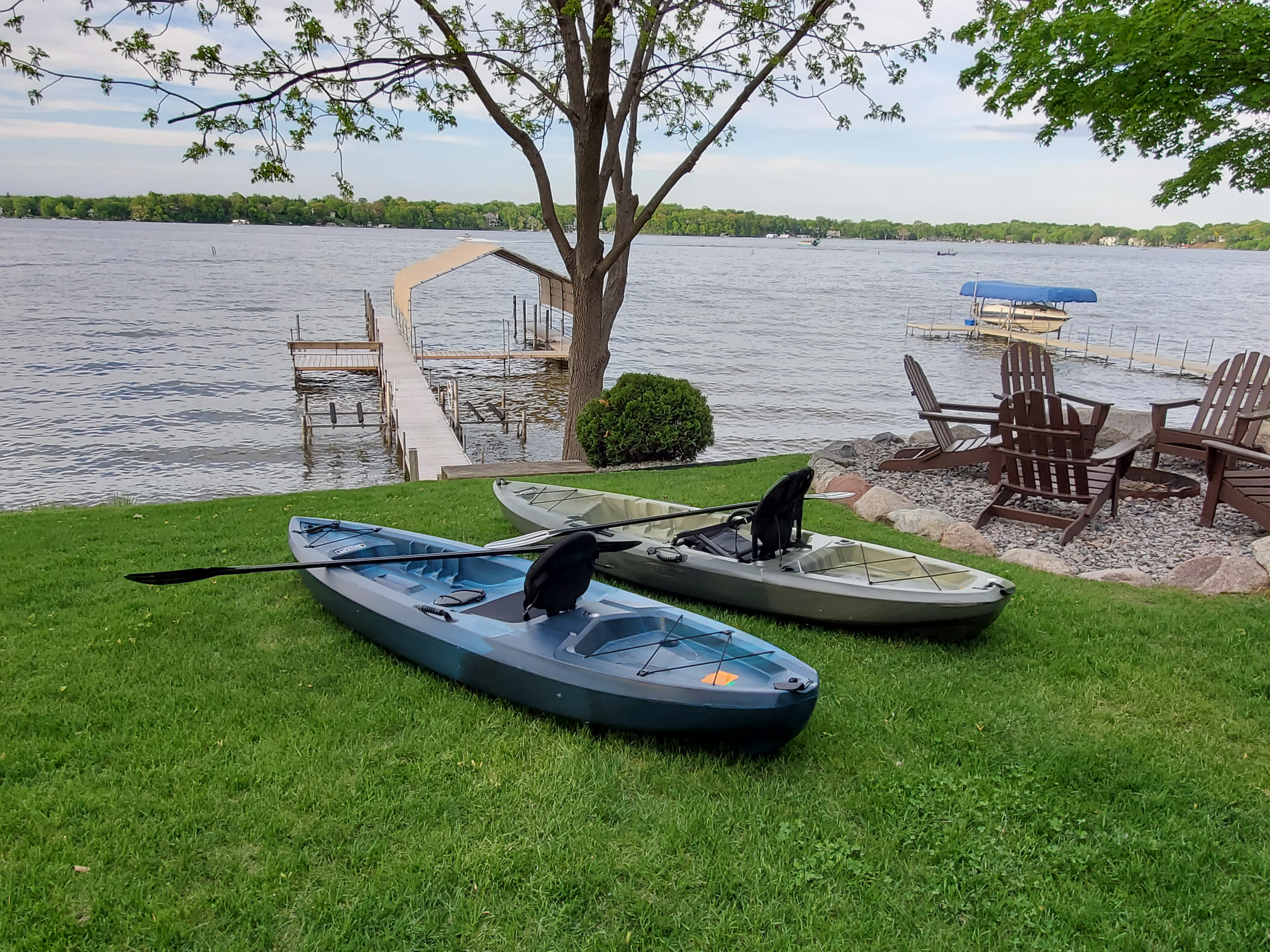 Two kayaks on a lakeside lawn with a dock and chairs in the background.