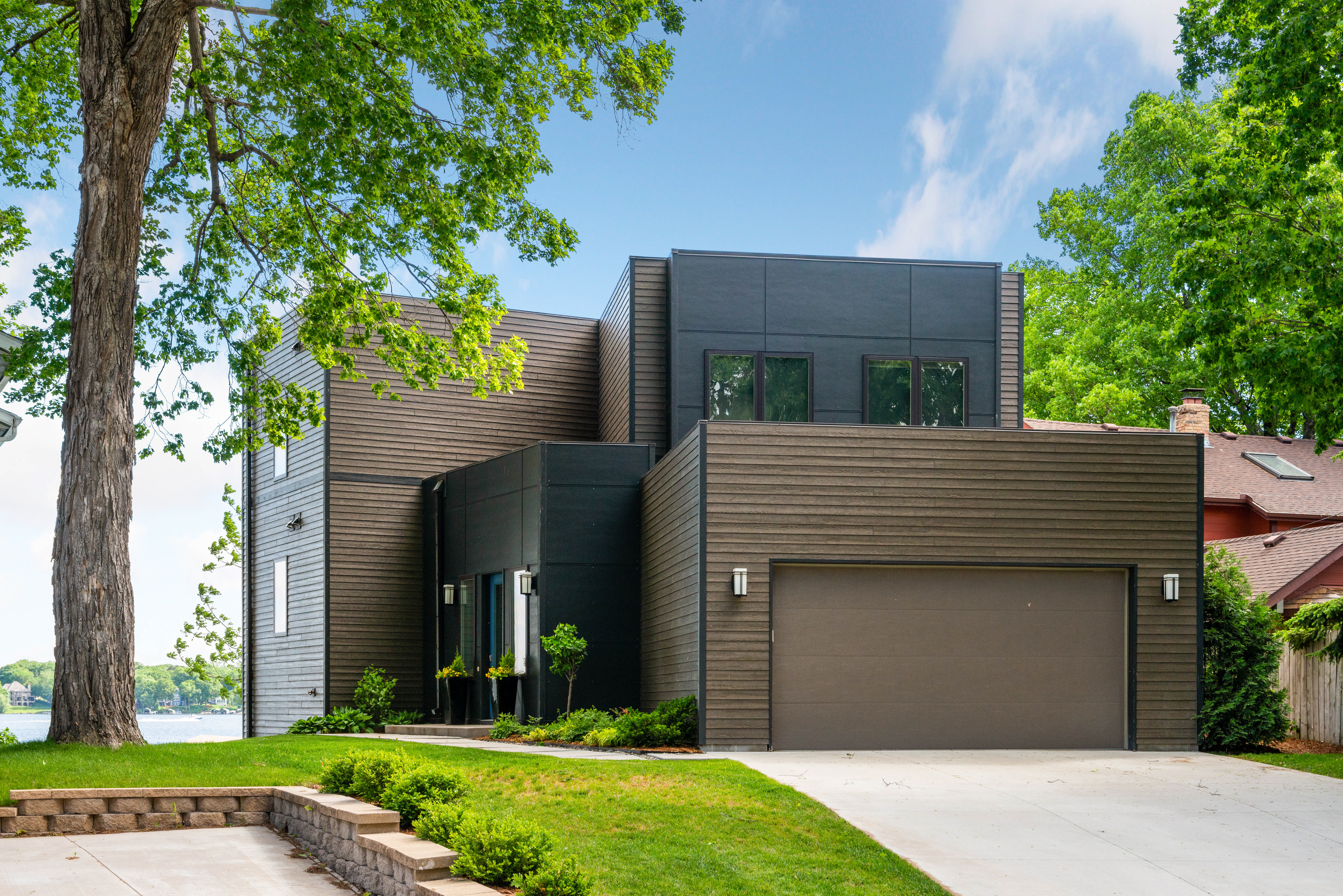 A modern two-story house with dark wood paneling in a serene lakeside setting.