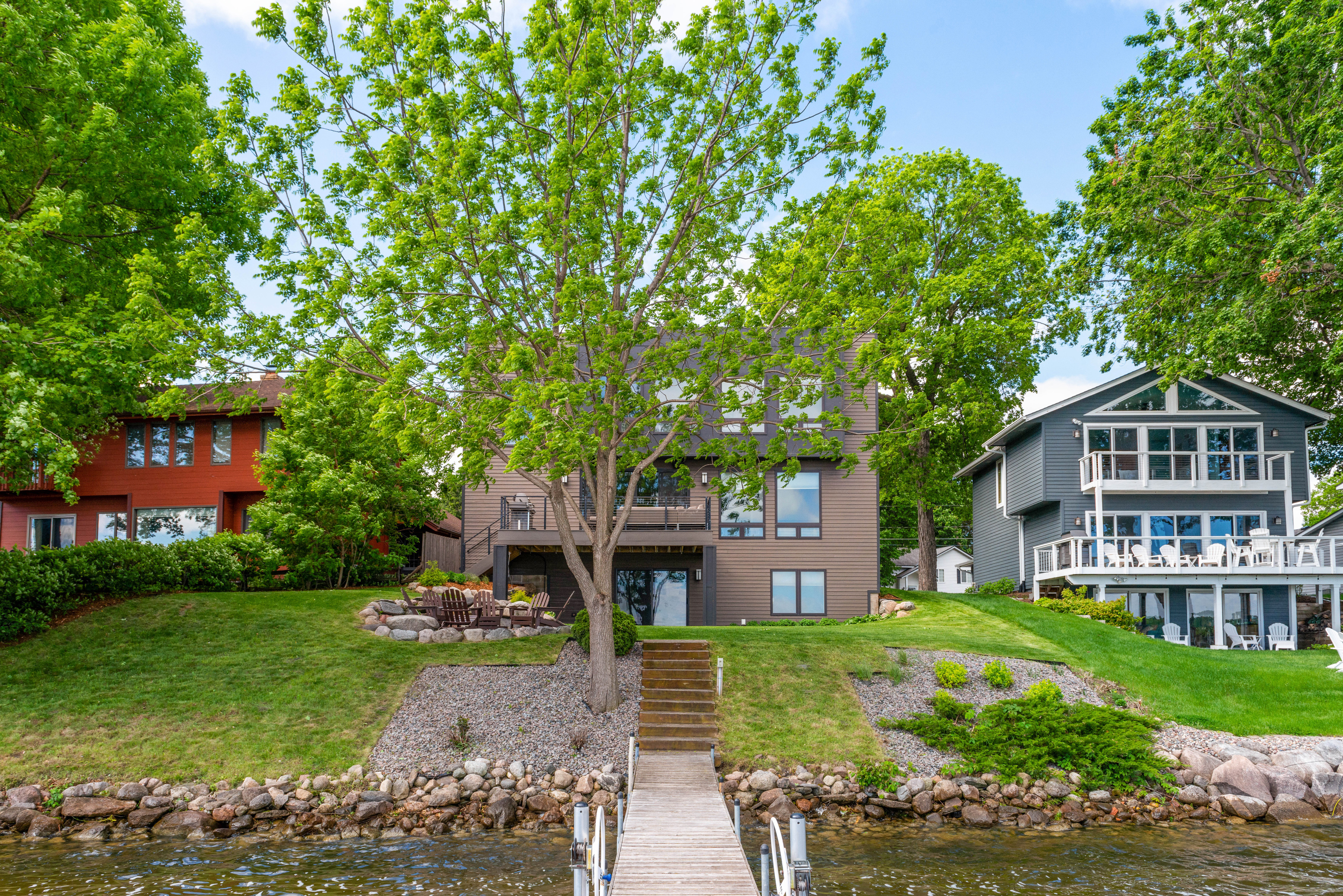 Lakeside homes surrounded by lush greenery, featuring a wooden dock and outdoor sitting area.