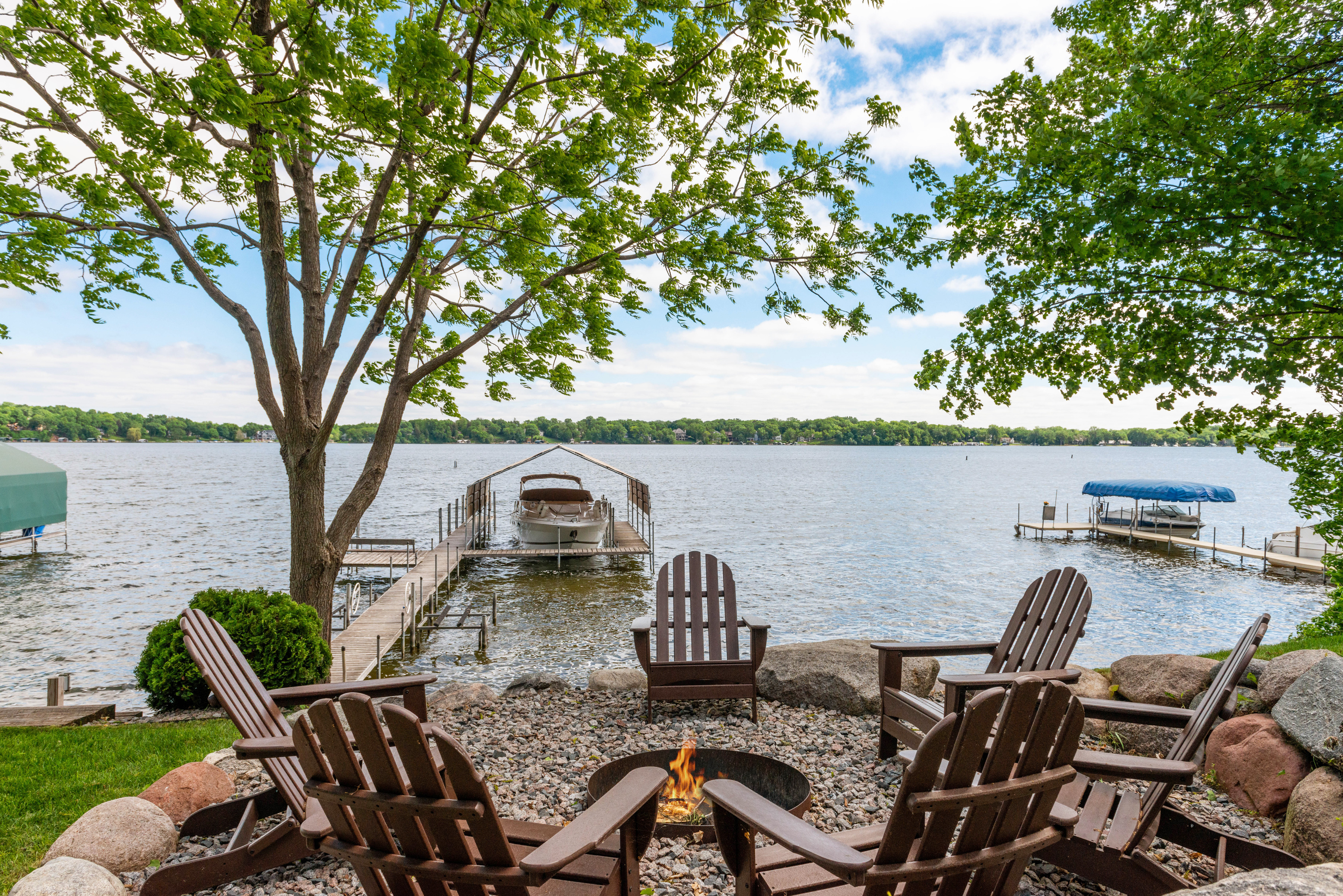 A cozy lakeside retreat with Adirondack chairs around a fire pit, overlooking a peaceful dock and boat.
