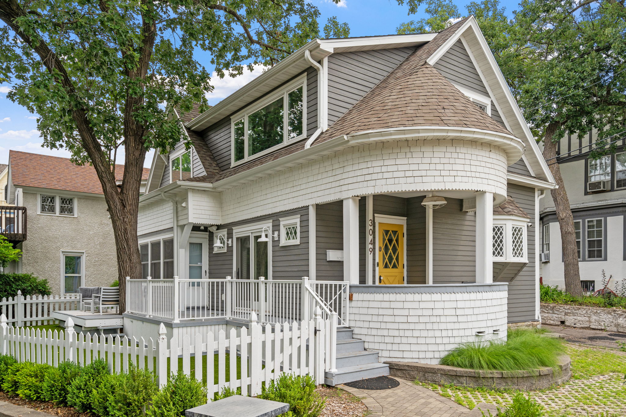 A gray and white house with a yellow door surrounded by greenery and a picket fence.