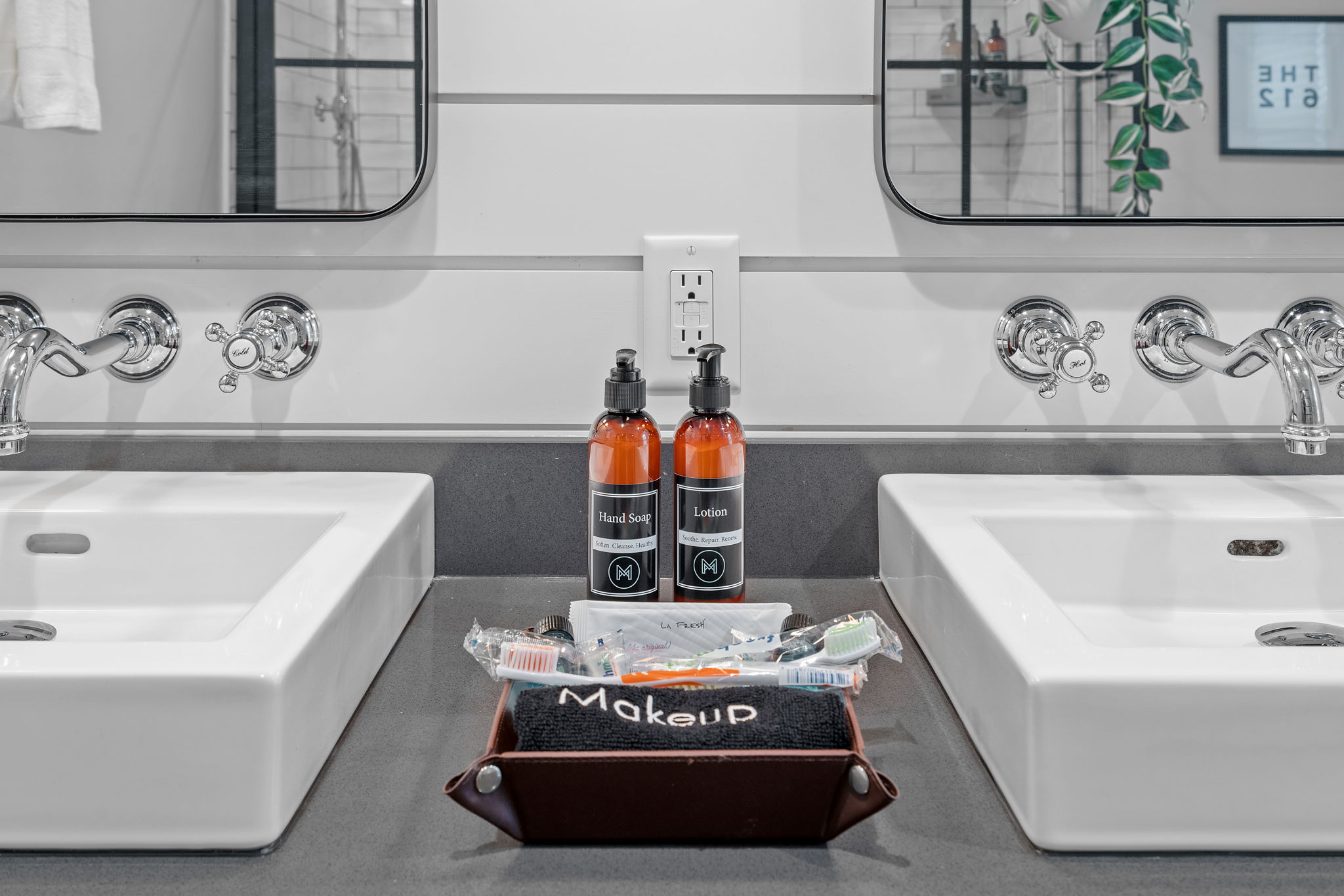 A modern bathroom countertop featuring two sinks, hand soap, lotion, and makeup essentials.