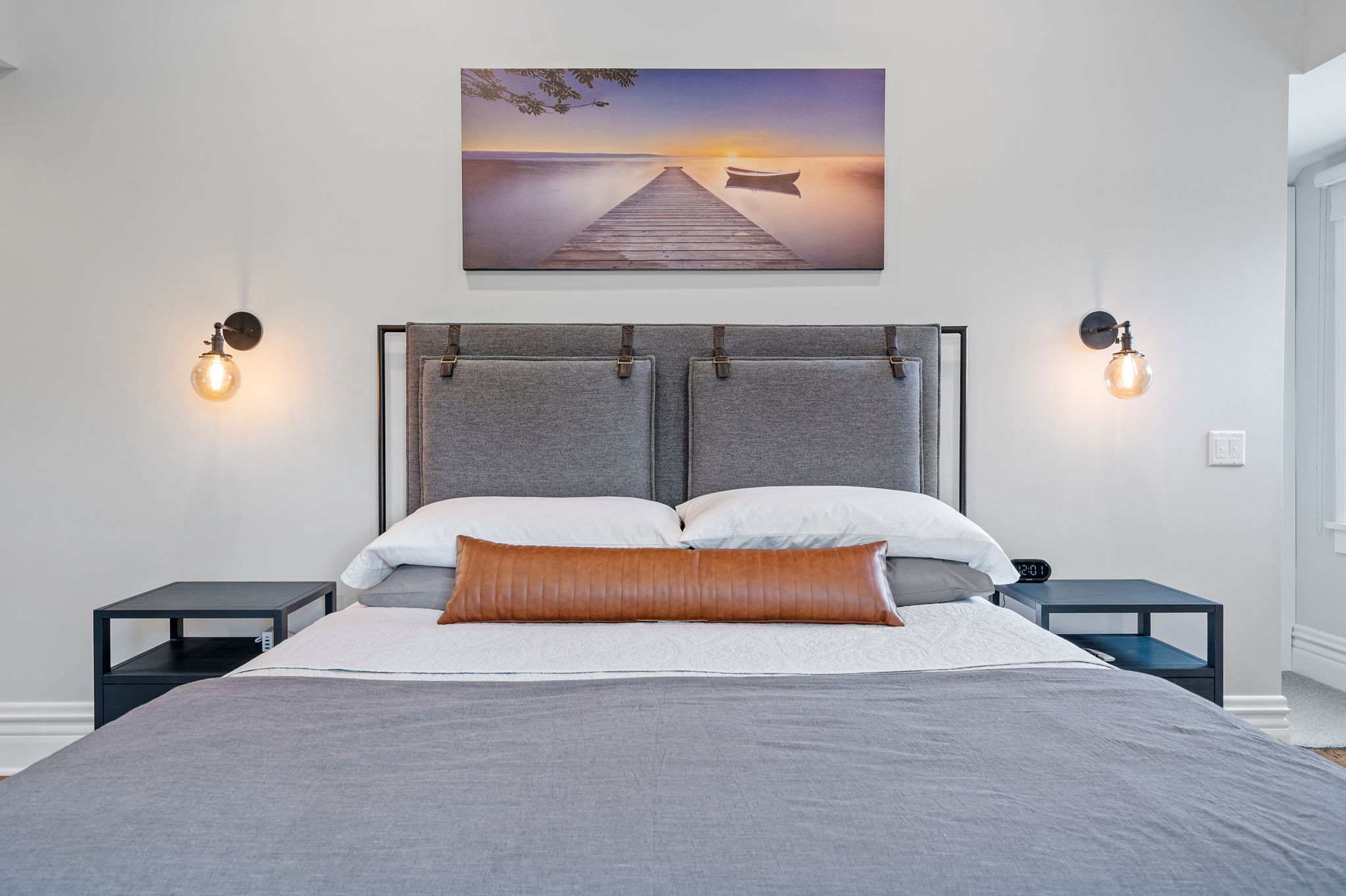 A modern and tranquil bedroom featuring a grey upholstered headboard, sunset lake photography, and ambient lighting.
