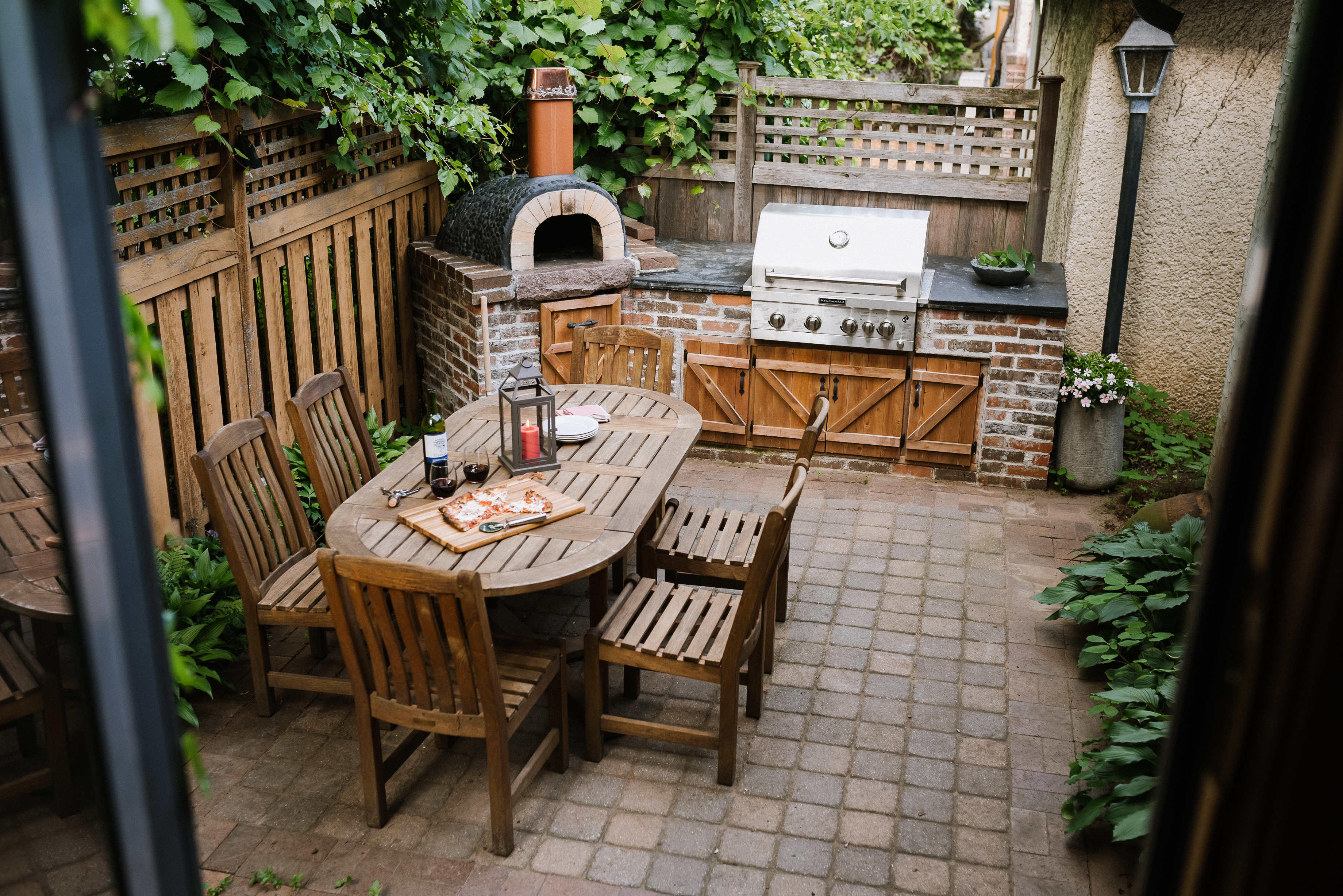 Cozy outdoor patio featuring a wooden table, chairs, and grill amidst greenery.
