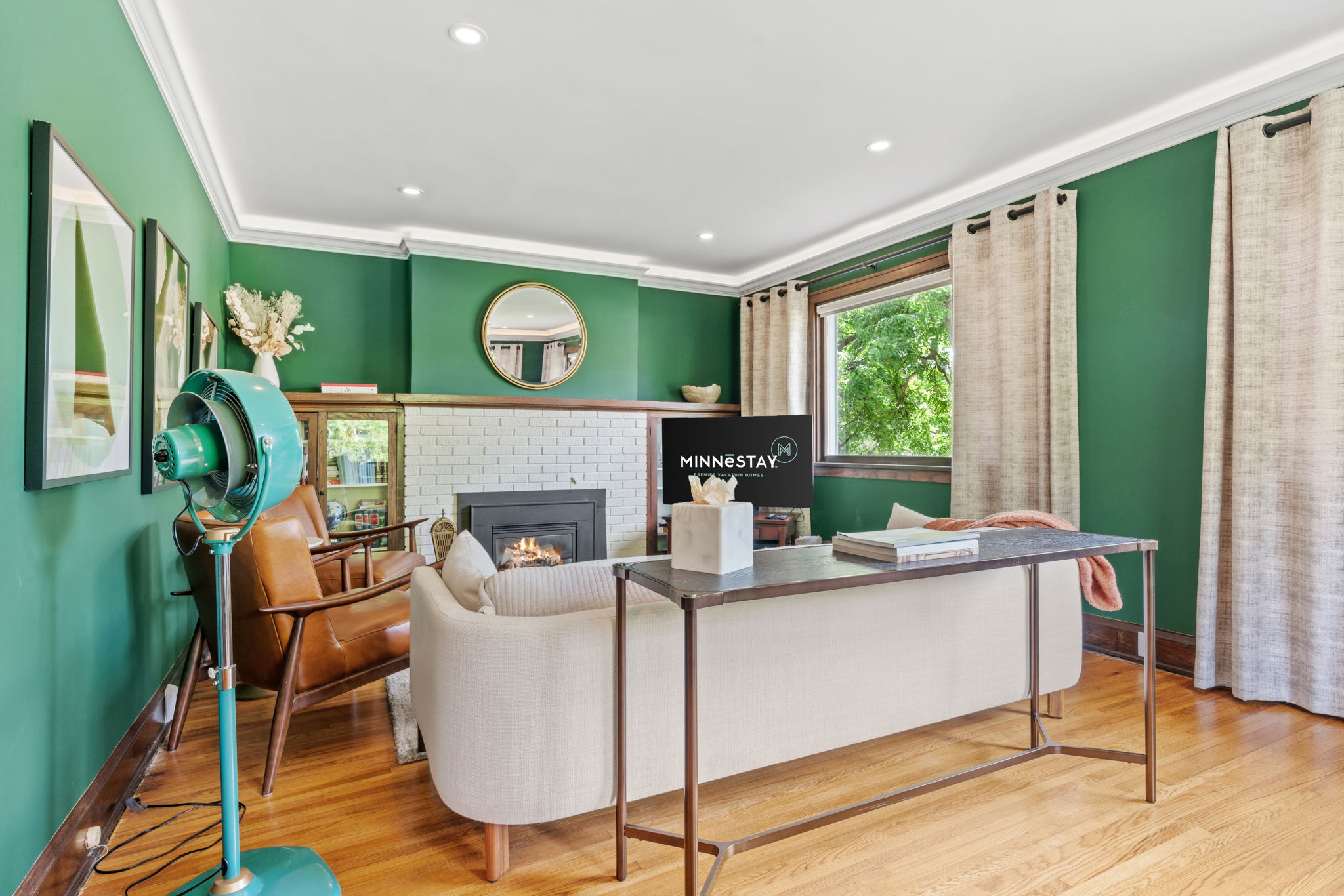 A modern living room with green walls, beige sofa, turquoise fan, and a white brick fireplace.