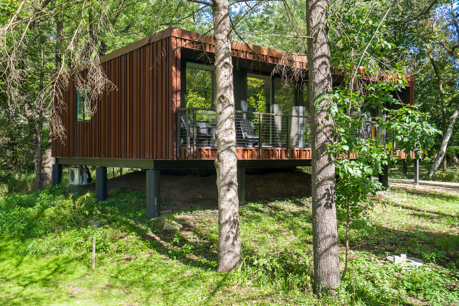 Modern elevated house with a rust-colored exterior and large windows, surrounded by trees and greenery.