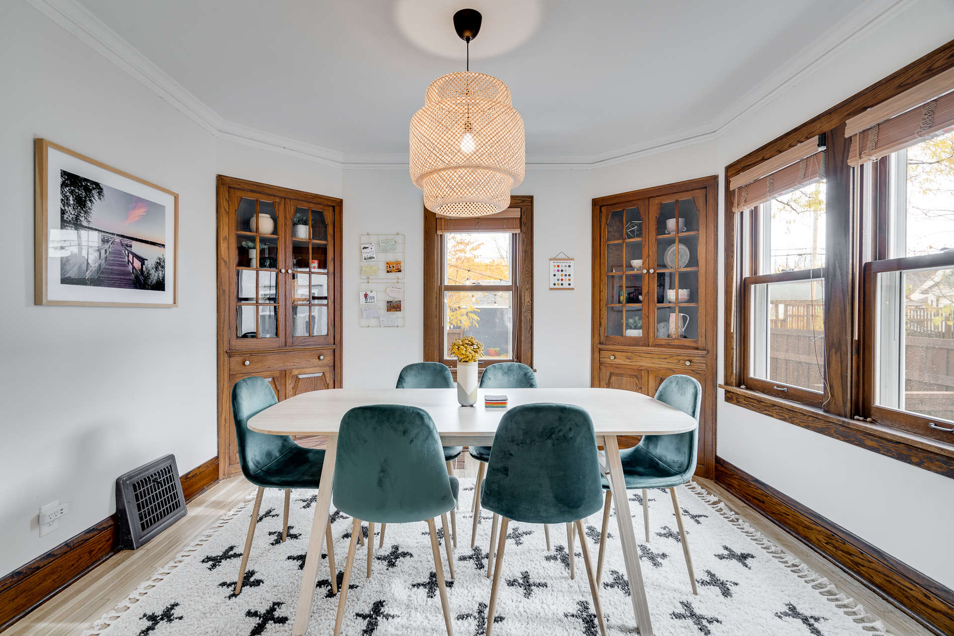 A modern dining room with an oval table, velvet chairs, and two wooden cabinets. A woven pendant light hangs above the table.