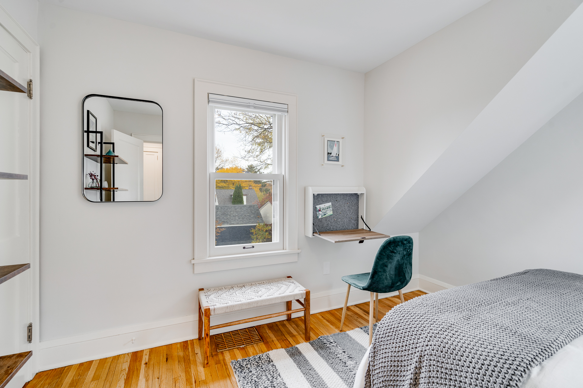 A minimalist room featuring a mirror, small desk, and bench with natural light streaming through the window.