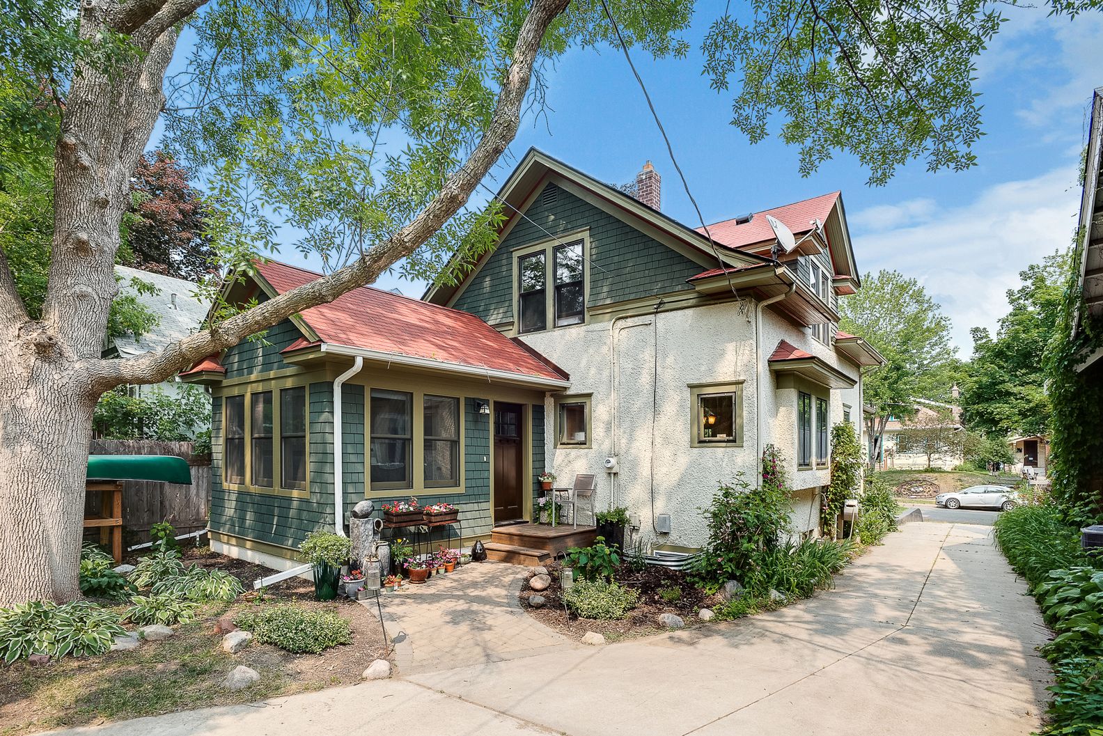 A charming two-story house with green shingles and a wooden porch, surrounded by lush greenery.