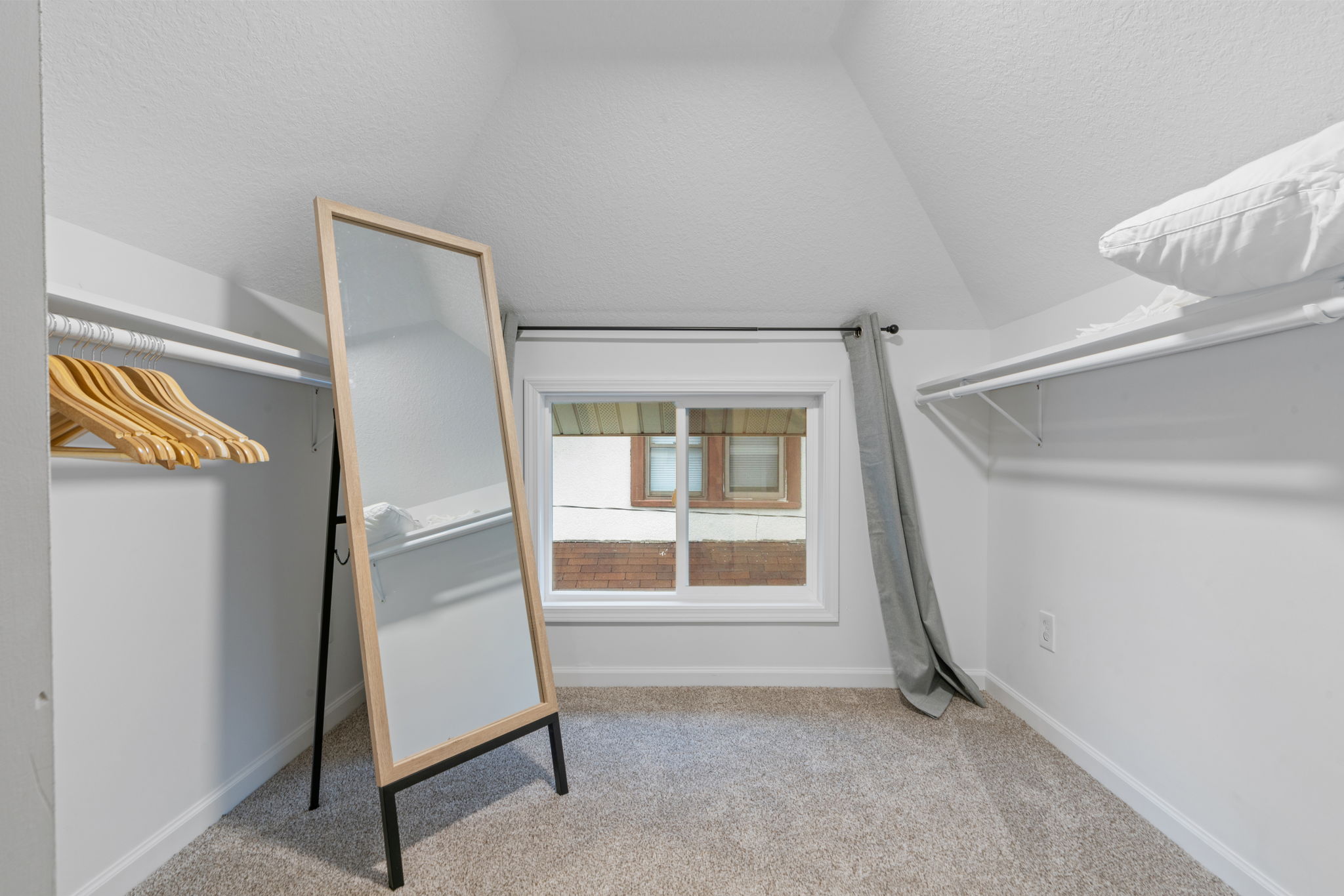 A cozy attic closet featuring wooden hangers, a framed mirror, and a window view.