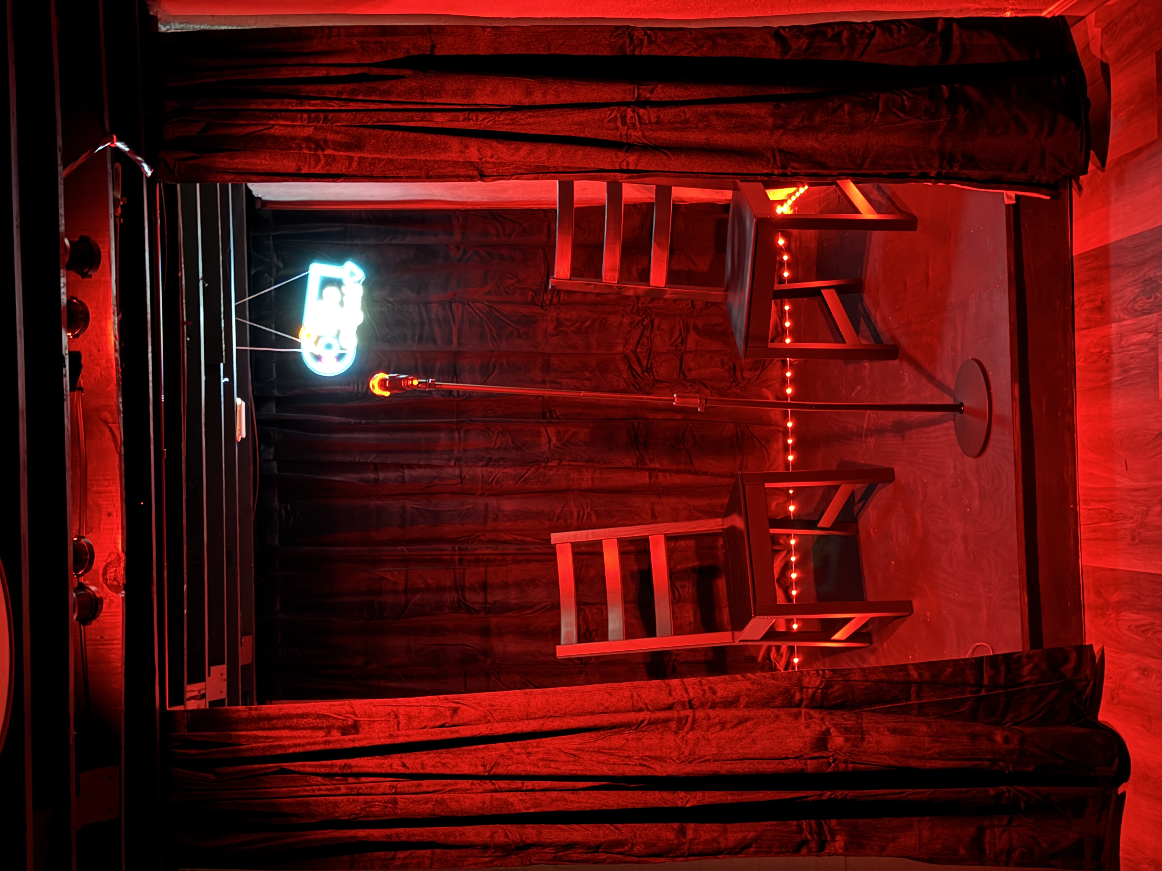 An intimate stage set for a performance with a microphone and two chairs, illuminated by red light and accented with neon and string lights.