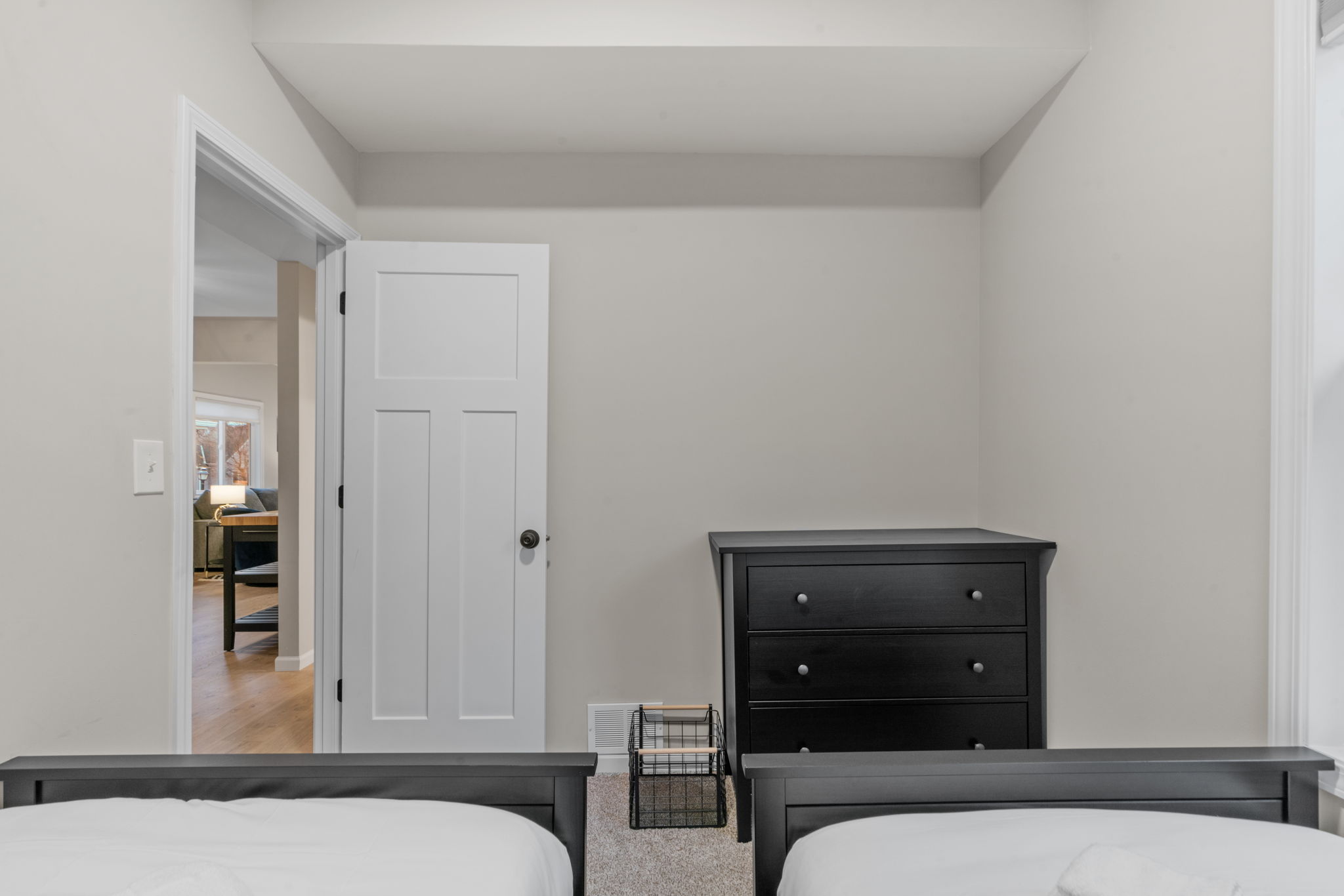 A minimalist bedroom featuring twin beds and an open door to a cozy living area.