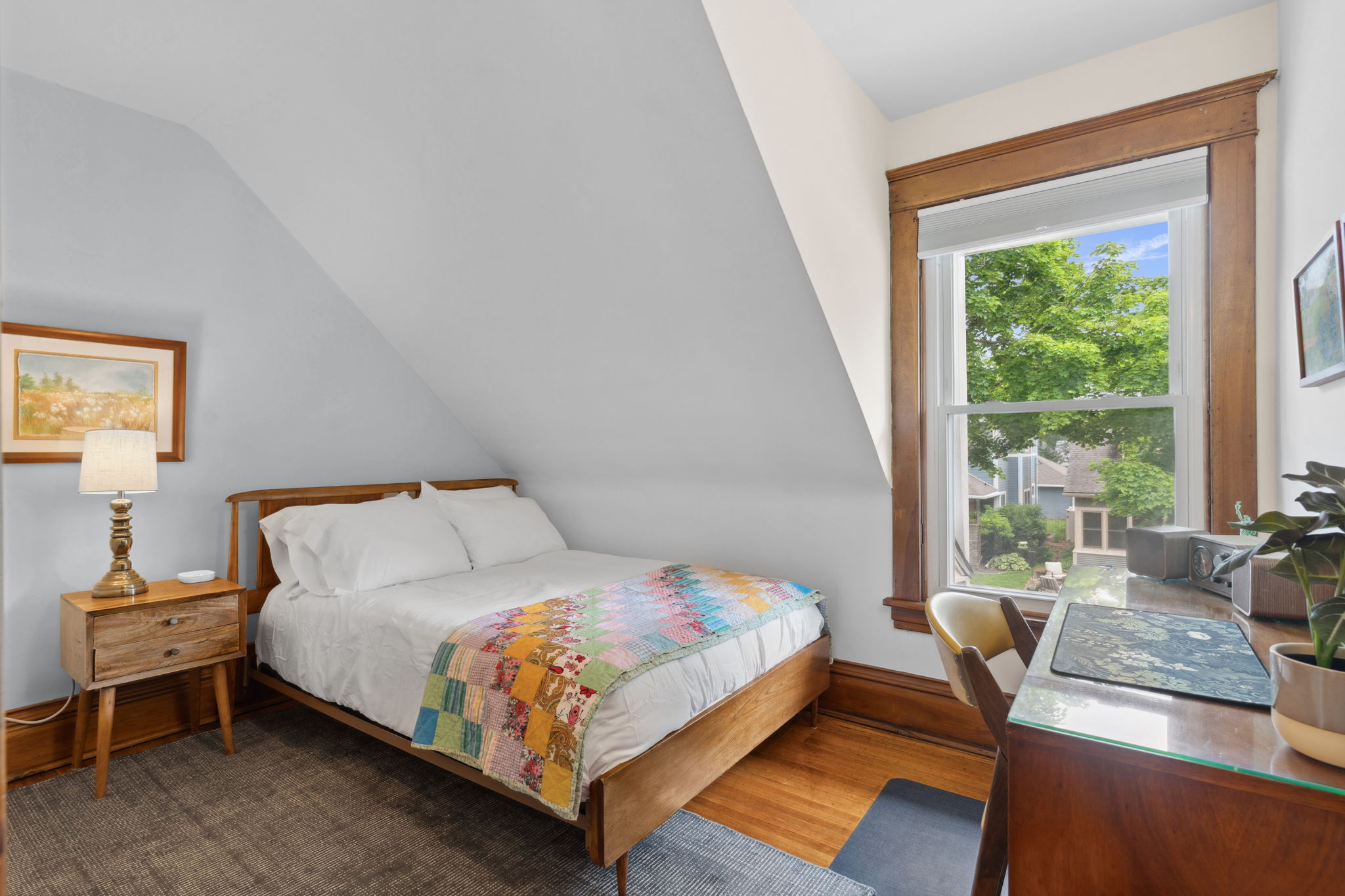A cozy bedroom with a sloped ceiling, featuring a colorful quilt and a sunny window view.
