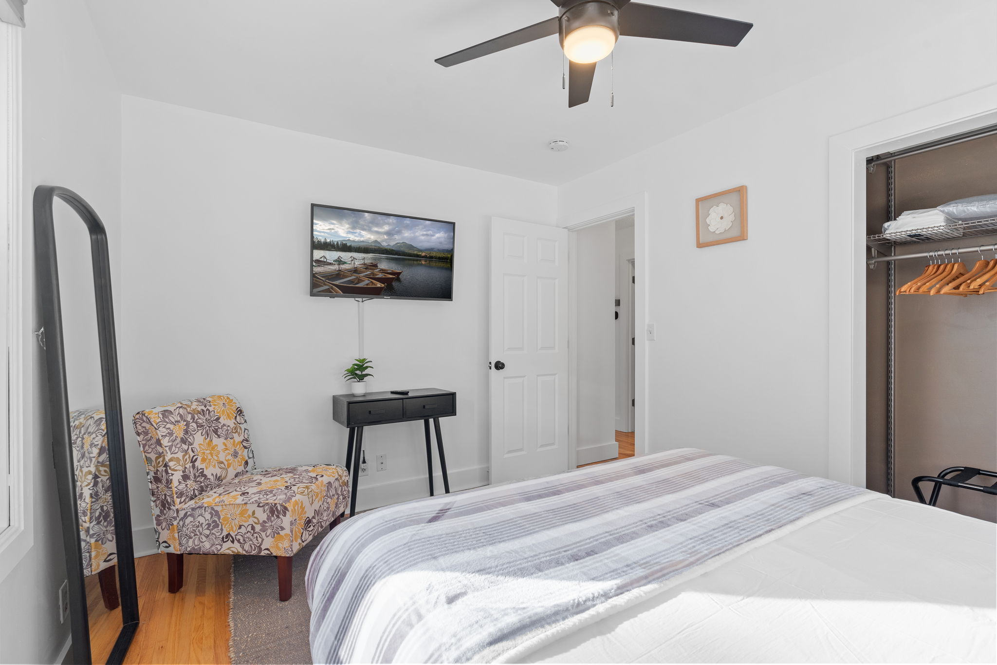A cozy, minimalist bedroom with a floral accent chair and a mounted TV displaying a serene lake view.