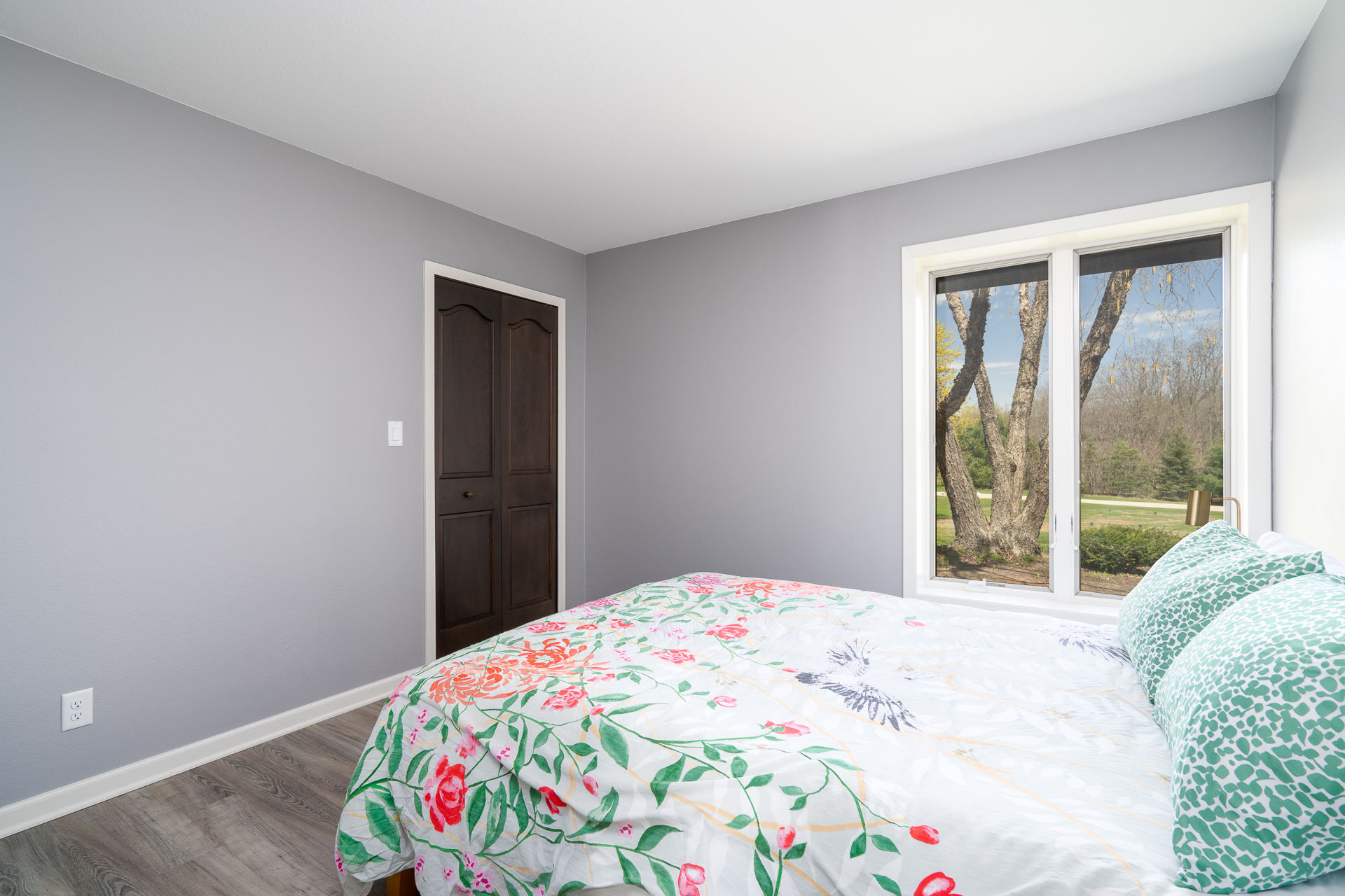 A cozy bedroom with floral bedding and a view of greenery outside.