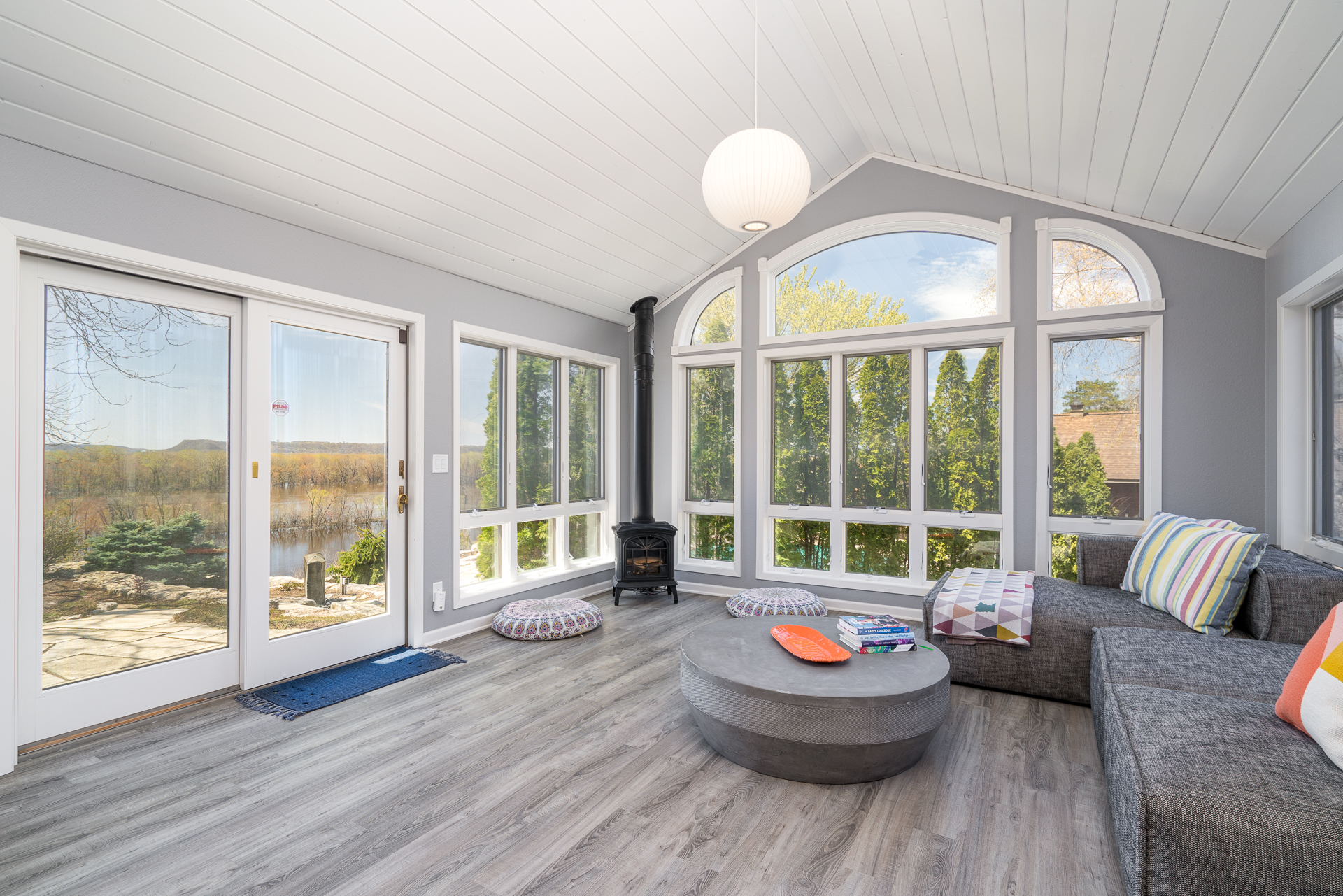 A cozy sunroom filled with light and vibrant furnishings, featuring large windows and a warm wood stove.