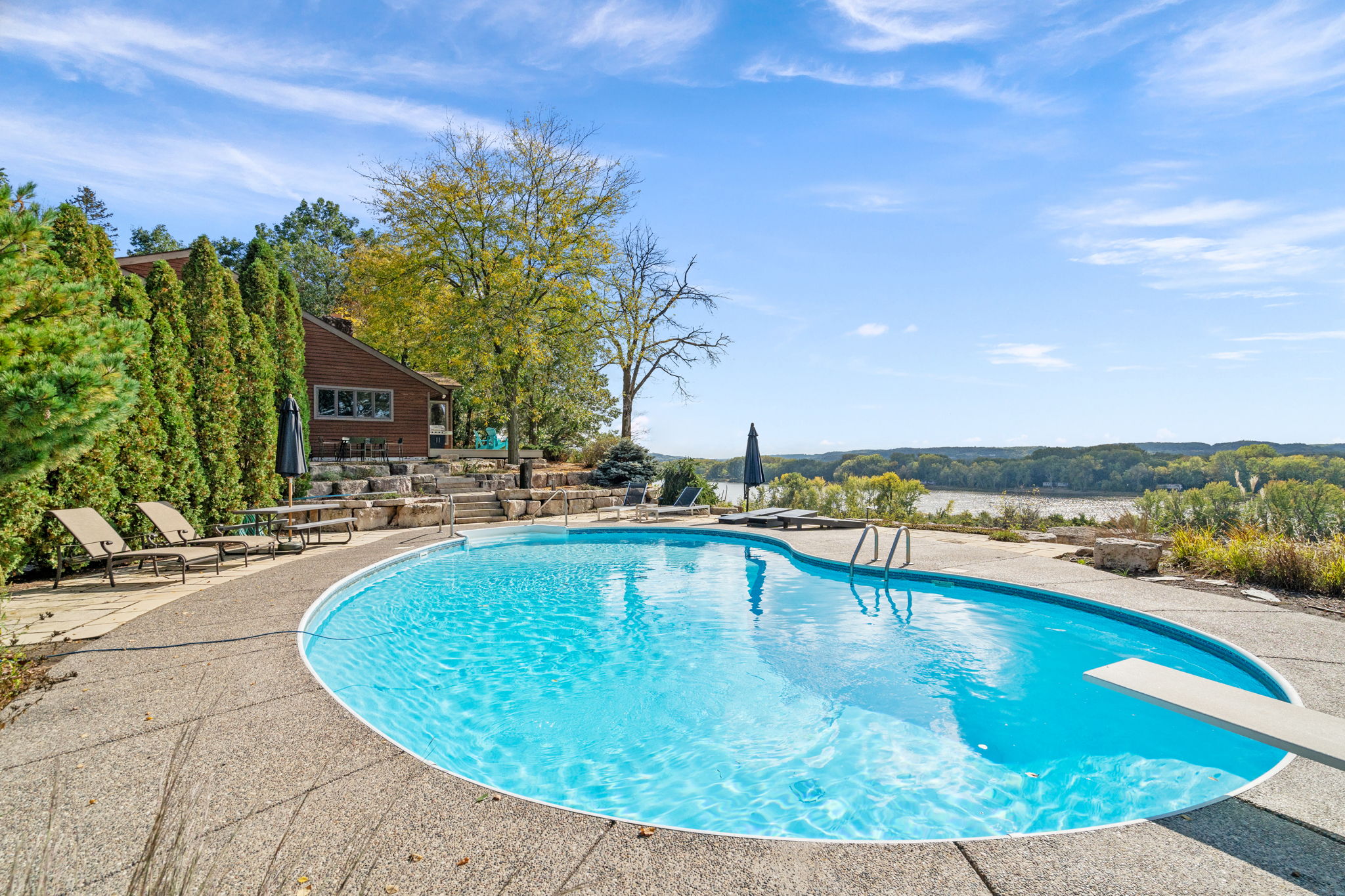 A tranquil backyard scene featuring a swimming pool overlooking a river and nestled amidst lush greenery.